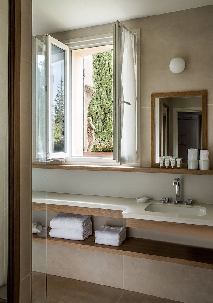 Natural light streams through open French windows into this serene bathroom, where warm wood accents and neutral tones create a spa-like sanctuary overlooking verdant gardens.