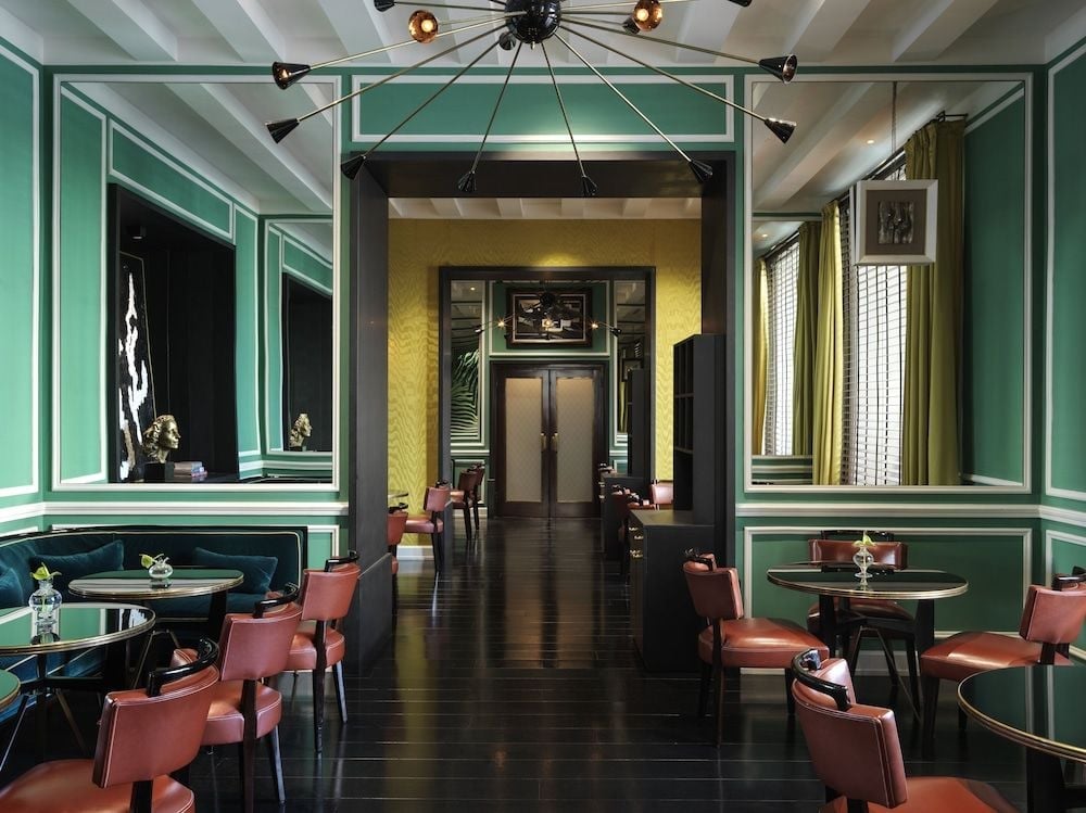 Emerald green paneling and cognac leather chairs create an intimate speakeasy atmosphere, where golden light from the statement chandelier casts shadows across polished dark floors.