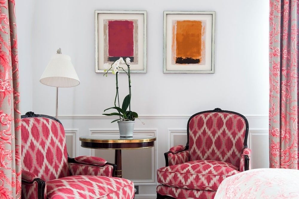Crimson ikat chairs create intimate conversation corners beneath abstract art, where morning light filters through rose-printed curtains in this elegantly appointed guest room.