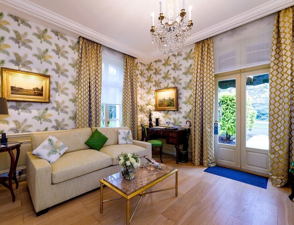 Palm-frond wallpaper and geometric curtains create sophisticated whimsy in this elegantly appointed suite, where crystal chandeliers cast warm light across honey-toned hardwood floors.