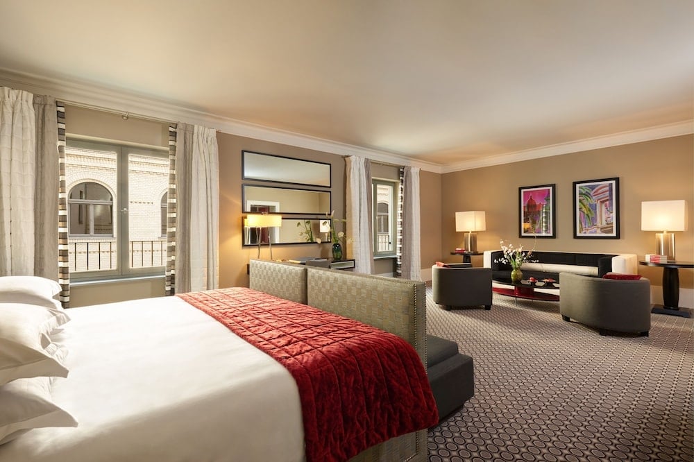 Warm taupe walls and geometric carpeting create sophisticated elegance in this spacious suite, where crimson velvet adds luxurious contrast to the refined neutral palette.