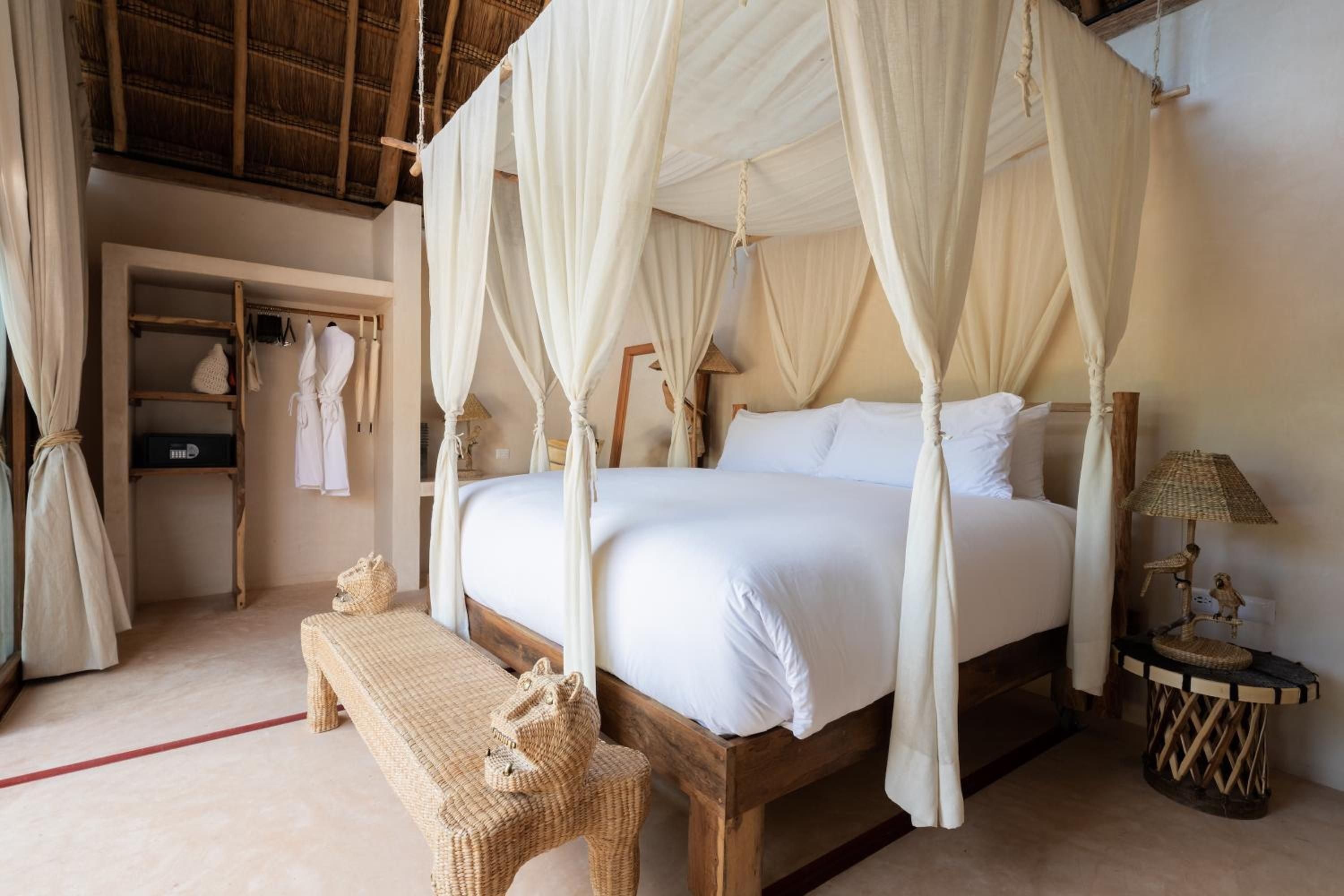 Flowing white linens frame a four-poster bed beneath rustic thatched ceilings, while woven textures and warm wood create an intimate jungle sanctuary.