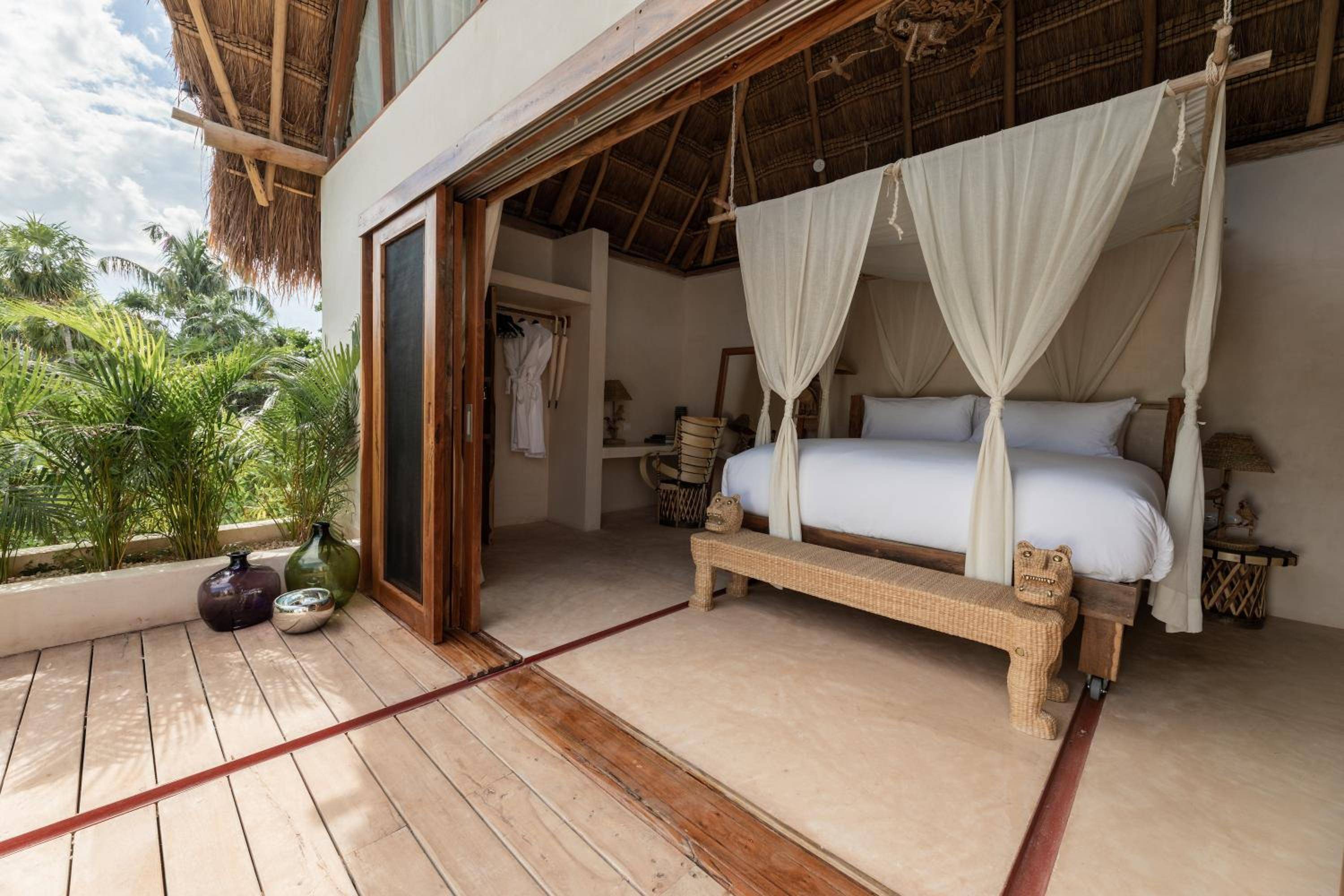 Flowing white curtains frame a four-poster bed beneath rustic palapa thatch, where polished concrete floors meet weathered wood decking overlooking jungle greenery.