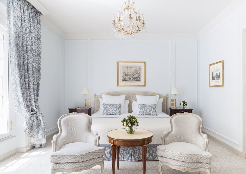 Powder-blue toile curtains frame tall windows while crystal chandeliers cast gentle light over cream bergère chairs and crisp white linens in this supremely elegant Parisian refuge.