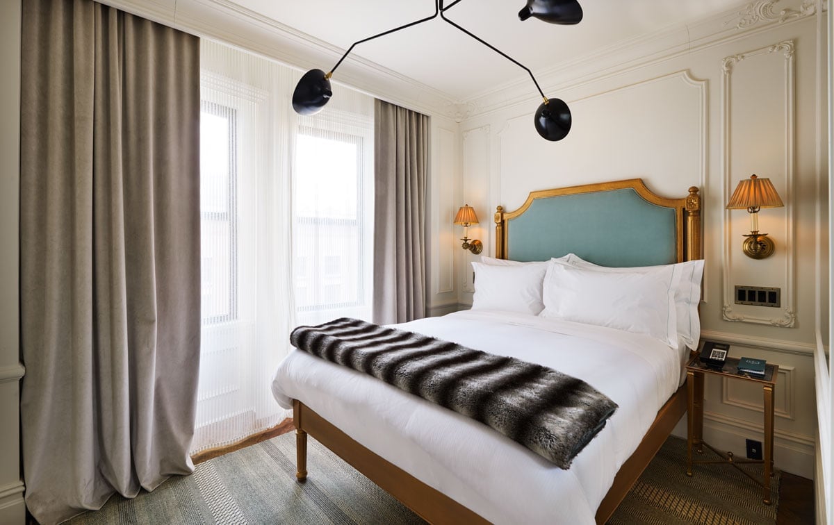 Crisp white linens meet sage velvet in rooms where modern black fixtures playfully contrast gilt-framed headboards and warm brass sconces.