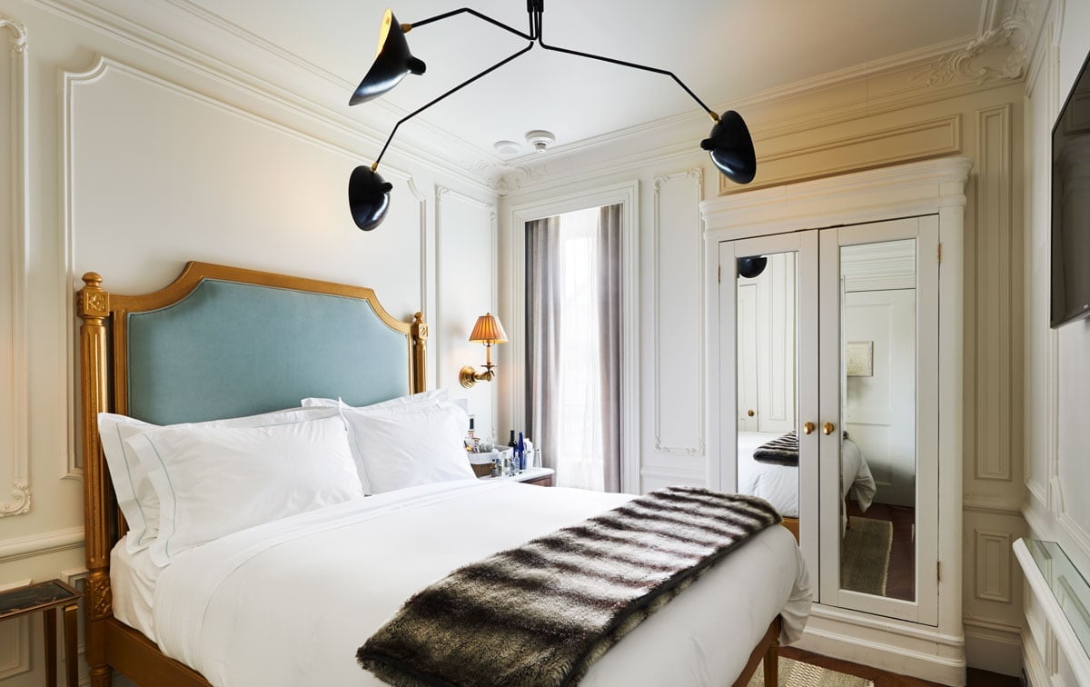 Parisian elegance meets Greenwich Village charm in this jewel-box room, where ornate moldings frame a peacock-blue headboard and modern Serge Mouille lighting creates intimate sophistication.
