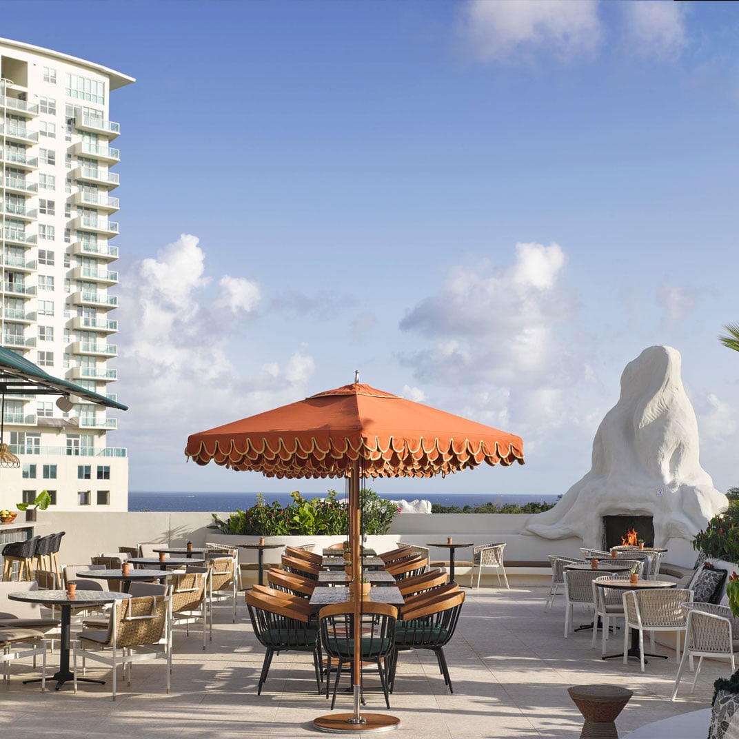 Orange-fringed umbrellas cast warm shadows across the rooftop terrace, where wicker chairs face both sculptural fire features and distant ocean views under Miami's endless blue sky.