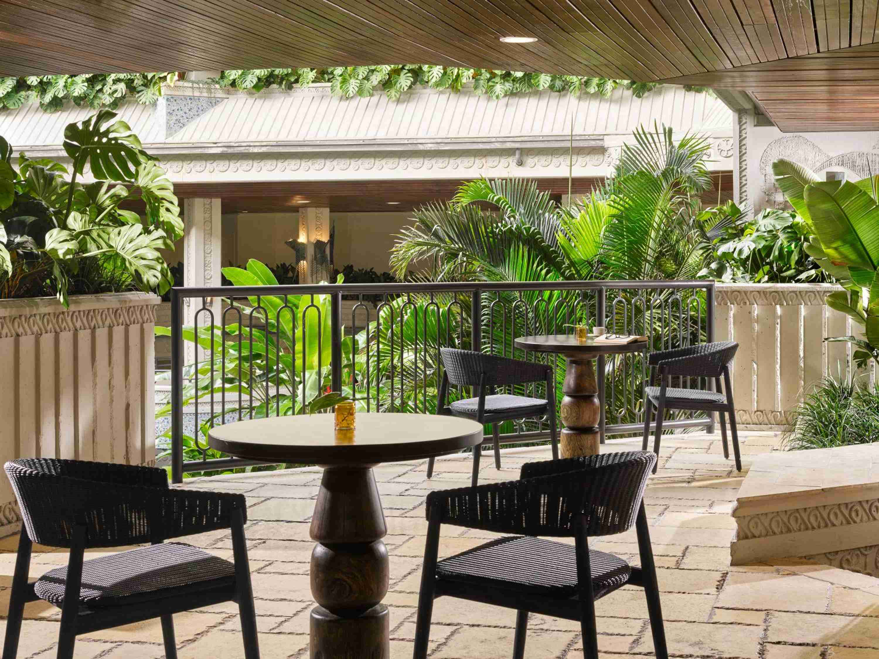 Wicker chairs and weathered wood tables create intimate dining nooks beneath cathedral ceilings, where tropical palms frame stone balustrades and dappled light filters through.