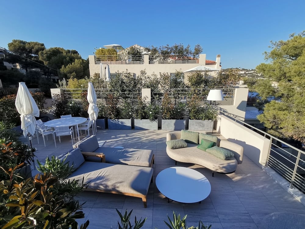 Curved lounge seating and scattered floor cushions create intimate conversation zones on this sun-drenched terrace, where Mediterranean light filters through olive branches.