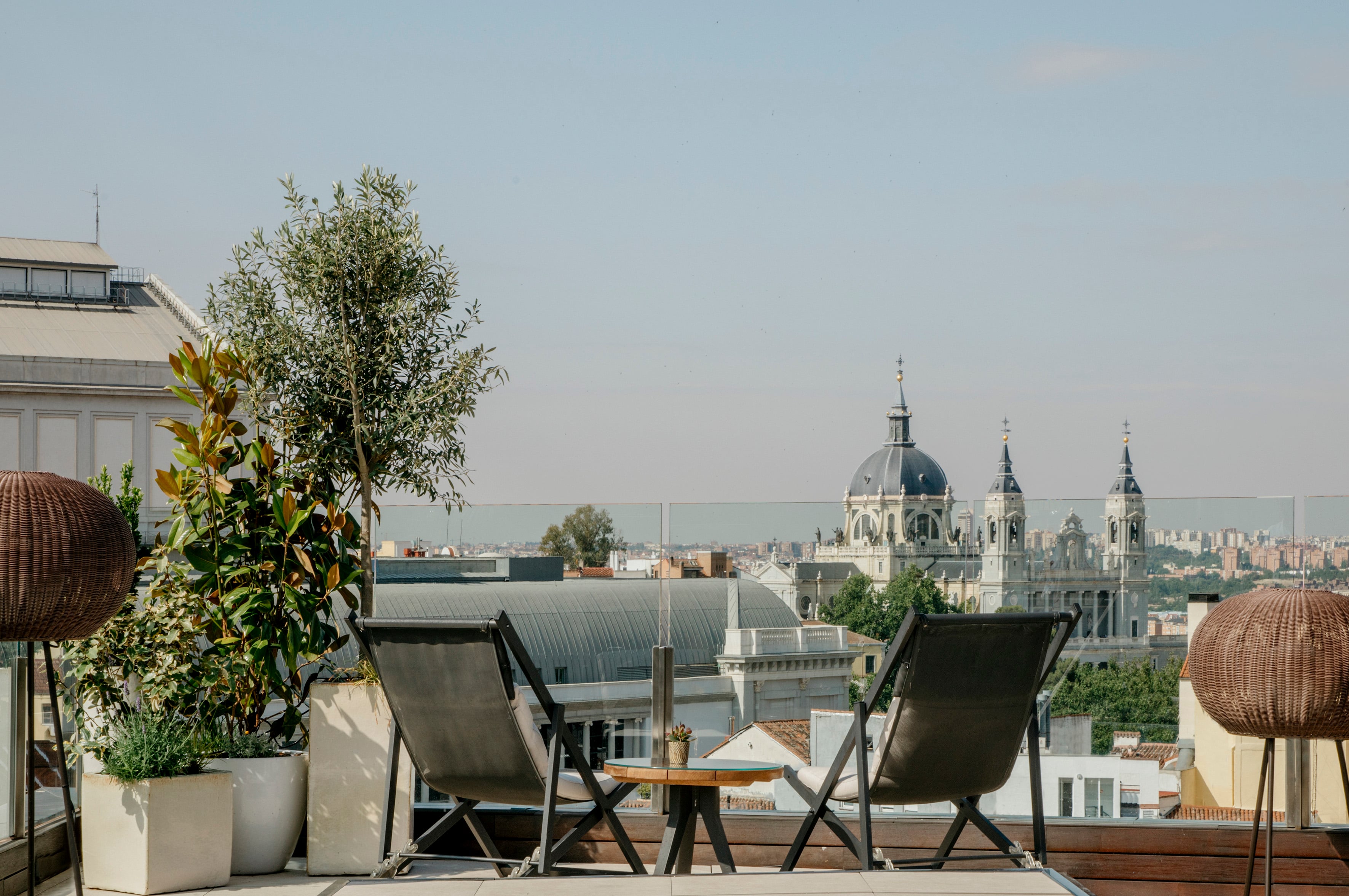 Wicker chairs frame Madrid's skyline from this elevated terrace, where potted olive trees catch golden light and cathedral domes punctuate the horizon.