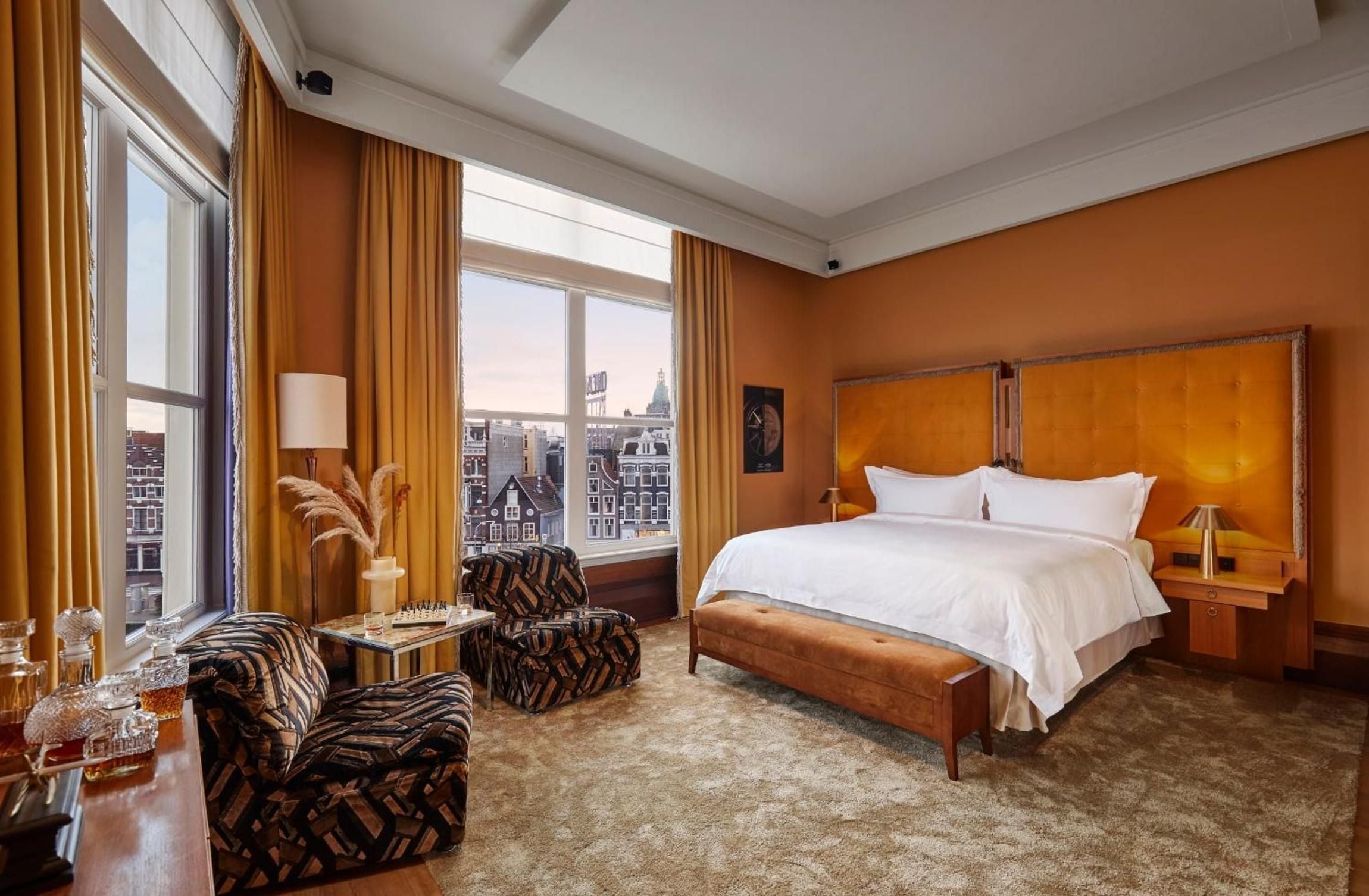 Warm amber walls and oversized windows frame Amsterdam's iconic canal houses, while geometric-patterned armchairs invite lingering over the cityscape beyond crisp white linens.