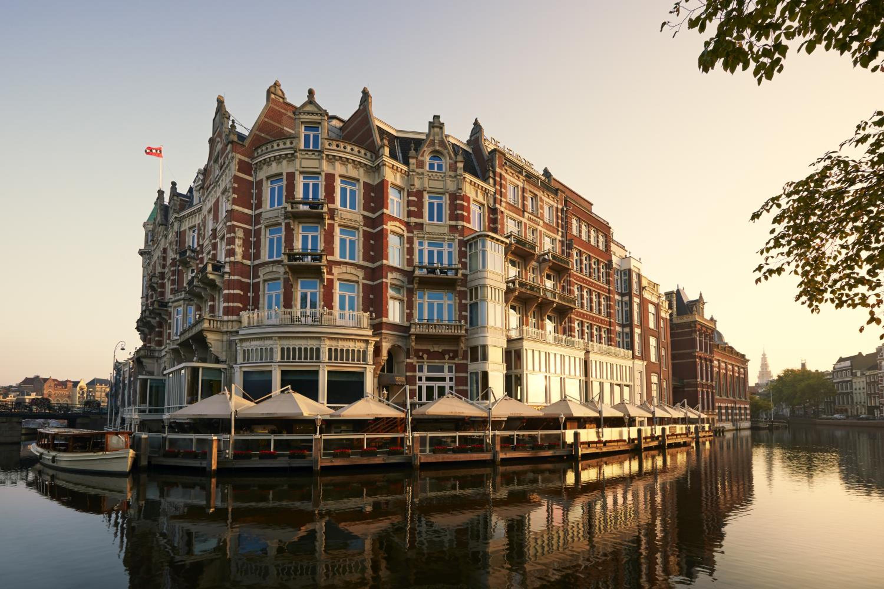 Golden hour light bathes the hotel's ornate Victorian facade as canal boats drift past, creating perfect reflections in Amsterdam's tranquil waterways.