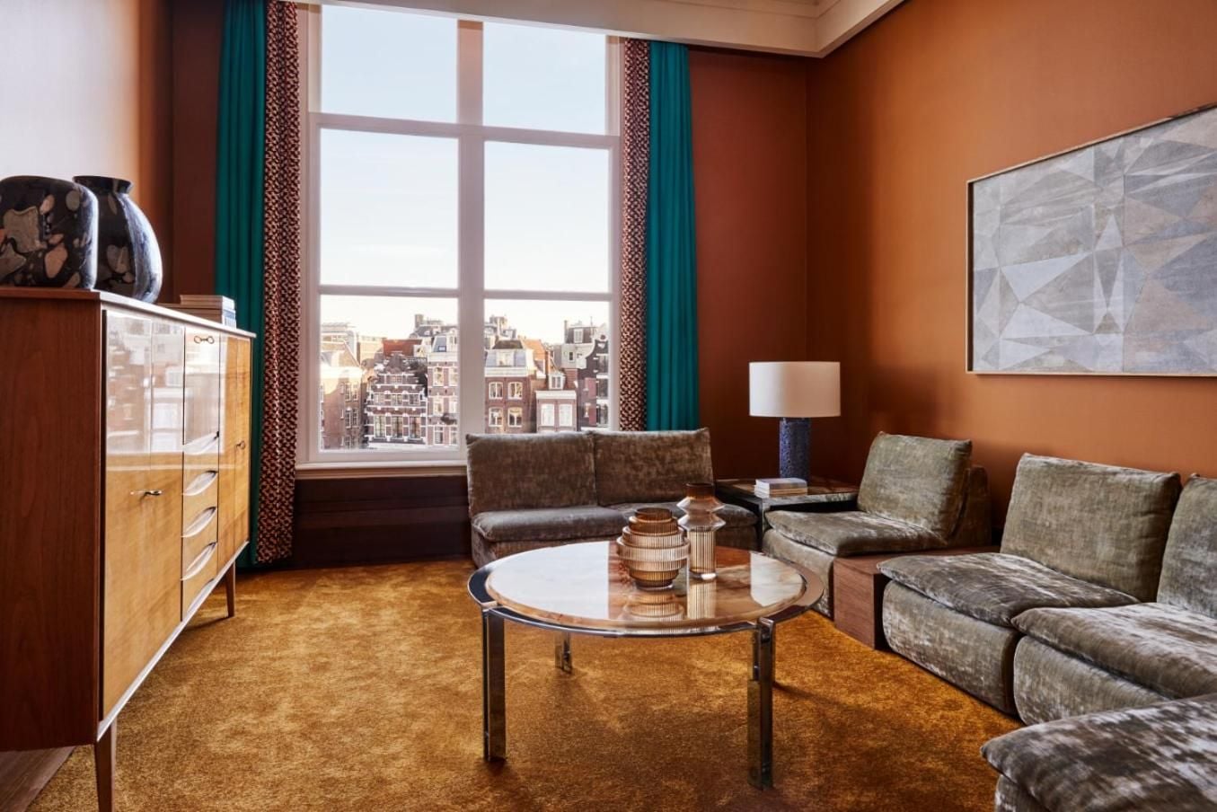 Warm terracotta walls and vintage velvet furnishings create an inviting retreat, while expansive windows frame Amsterdam's iconic canal-side architecture in golden afternoon light.