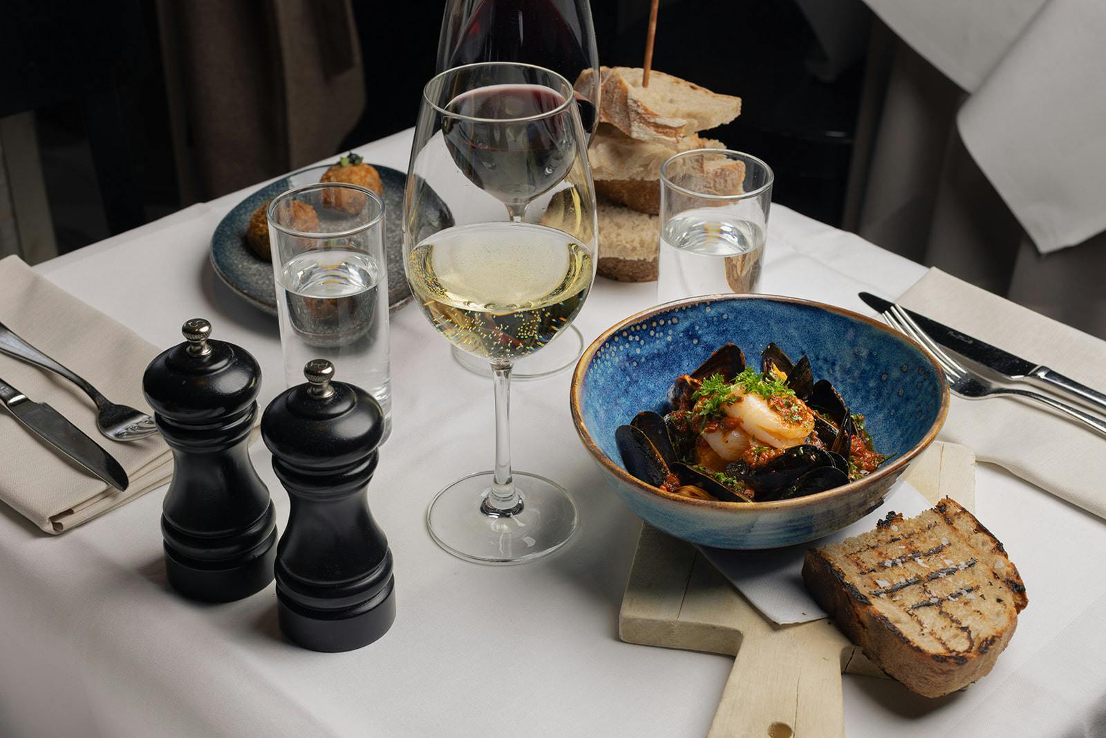 Artfully plated mussels nestle in a cobalt ceramic bowl while white wine catches afternoon light—Nordic culinary artistry meets intimate dining sophistication.