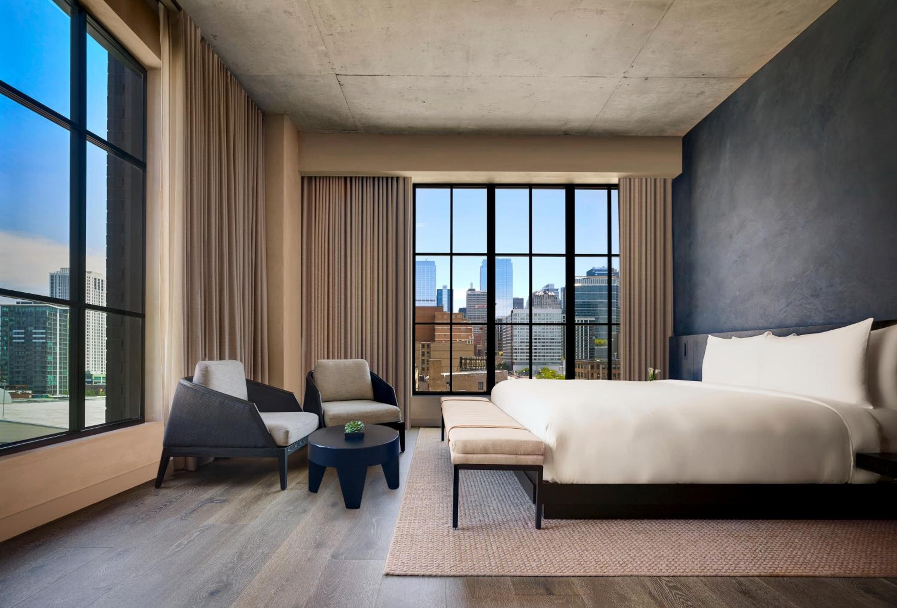 Floor-to-ceiling windows frame Chicago's skyline from this corner suite, where exposed concrete meets warm taupe curtains and crisp white linens invite urban sanctuary.