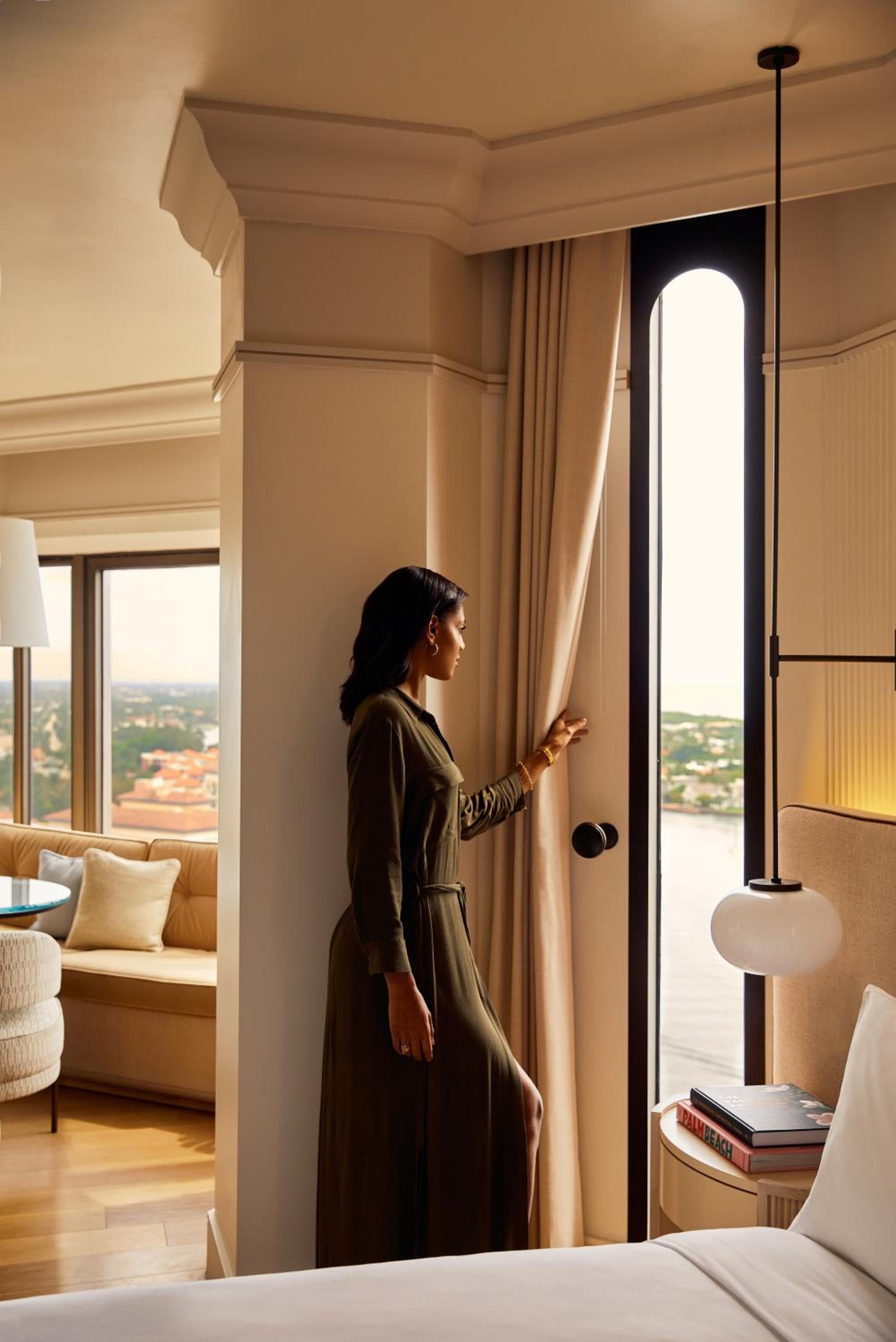 Warm afternoon light filters through floor-to-ceiling windows as a guest draws back curtains to reveal sweeping views of Boca Raton's coastline from this elegantly appointed tower suite.