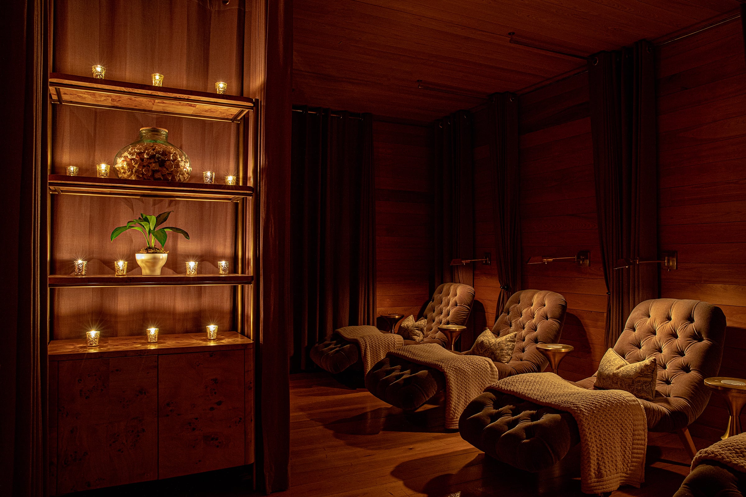 Amber-lit relaxation loungers cocoon guests in tufted luxury, while flickering votive candles and warm wood create an intimate sanctuary for pre-treatment tranquility.