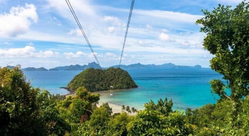 Zip lines slice through tropical canopy toward El Nido's pristine coastline, where turquoise waters cradle forested islets beneath scattered clouds.