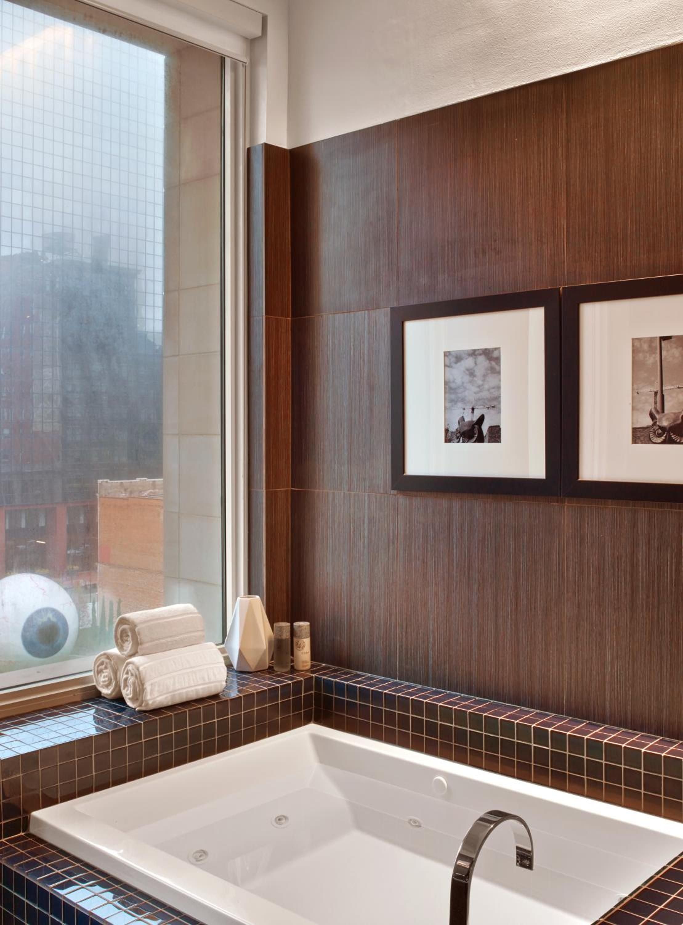 Rich walnut paneling and chocolate mosaic tiles frame the soaking tub, where rolled towels and city views create an urban spa sanctuary high above Dallas.