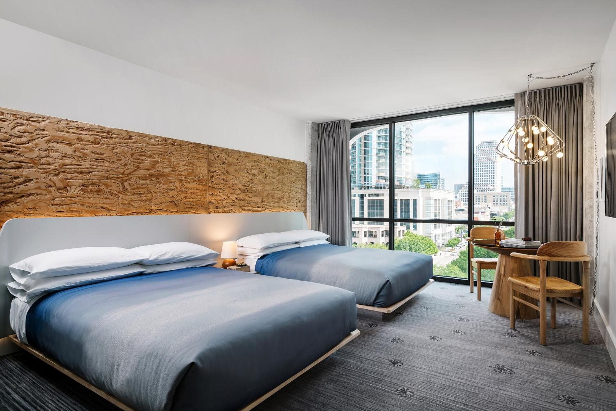 Twin beds with gradient blue linens face floor-to-ceiling windows showcasing Austin's skyline, while a striking wood-carved headboard adds organic warmth to the contemporary space.
