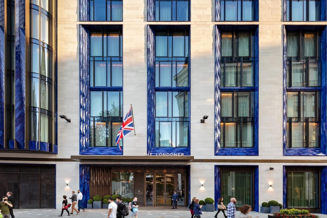 Blue-framed windows create striking geometric patterns across the limestone facade, while the Union Jack proudly marks this sophisticated Leicester Square sanctuary amid London's bustling crowds.