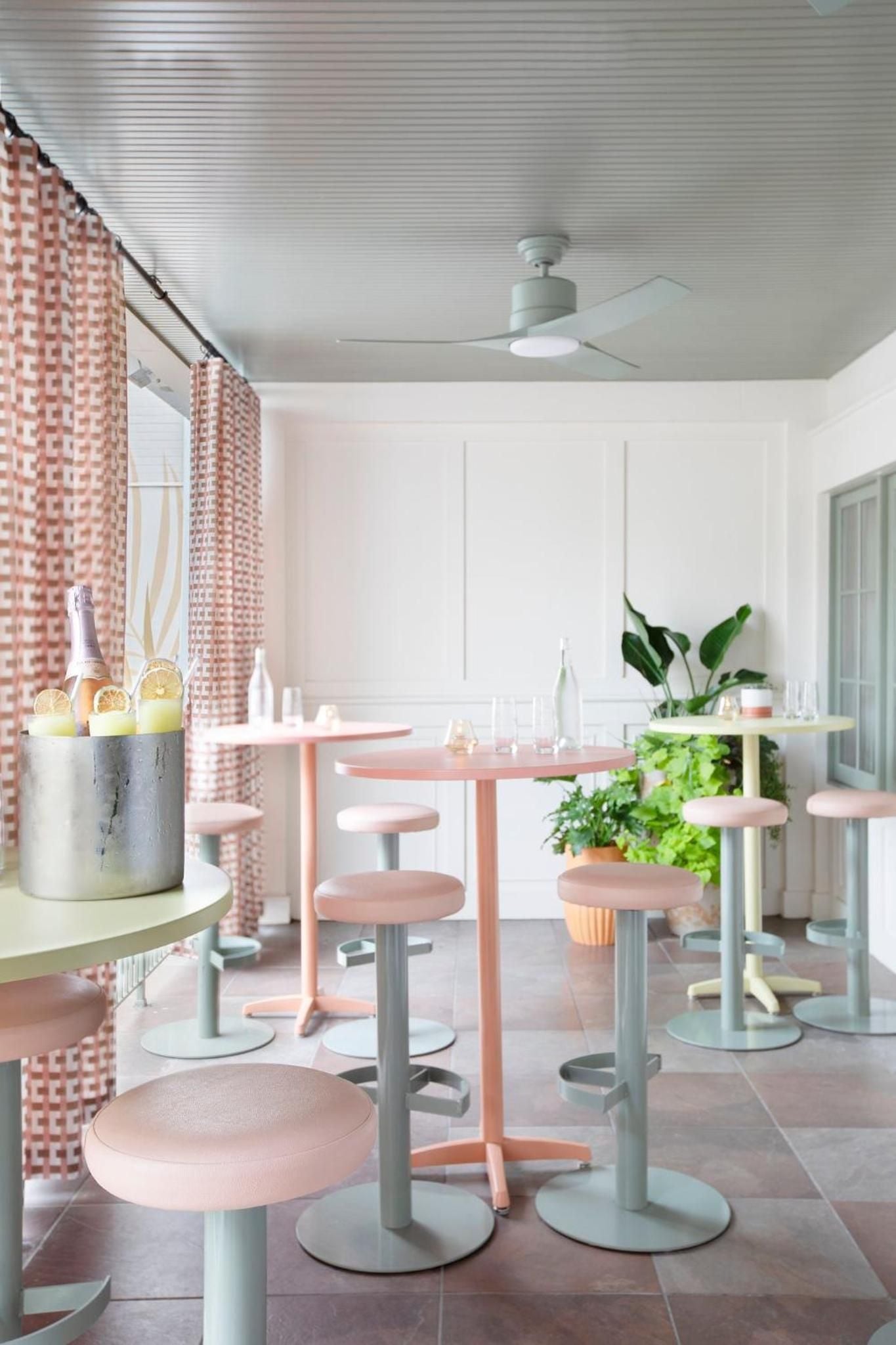 Coral-hued bar stools gather around peachy tables beneath corrugated ceilings, where craft cocktails chill in marble buckets amid trailing greenery and soft industrial charm.