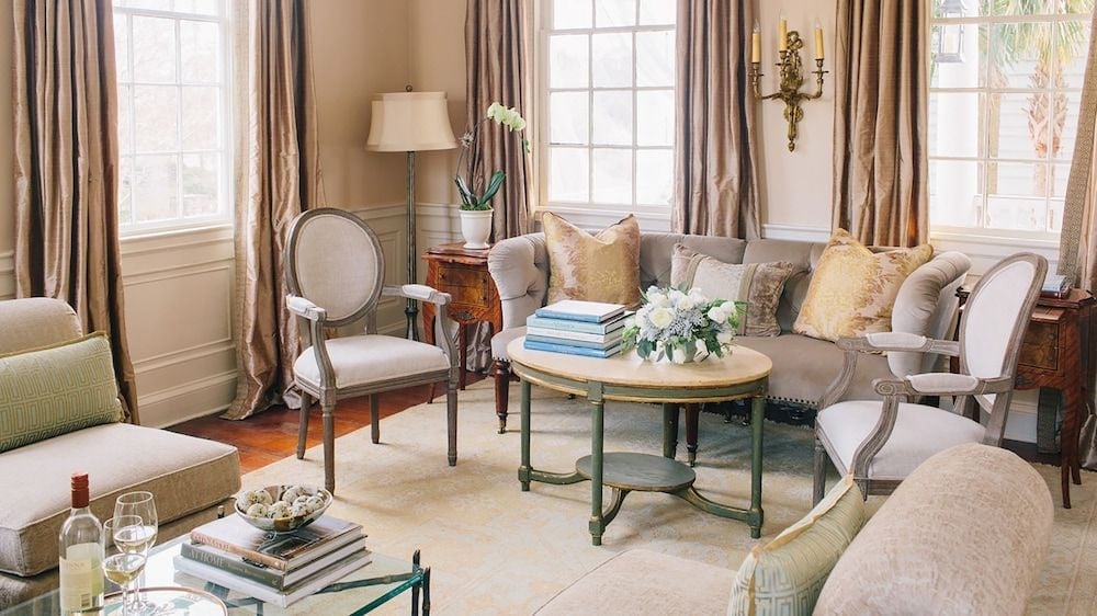 Soft afternoon light filters through elegant curtains in this sophisticated sitting room, where neutral linens meet antique wood and fresh orchids suggest refined Southern hospitality.