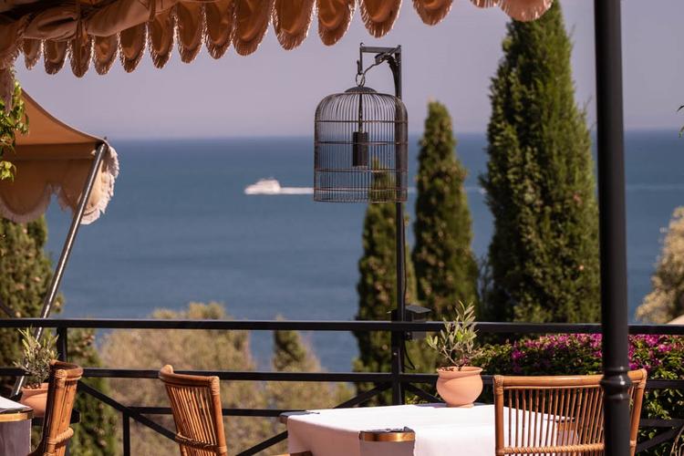 Wicker chairs frame intimate terrace dining where a vintage birdcage catches Mediterranean light, Cyprus trees standing sentinel over the sparkling Tyrrhenian Sea below.