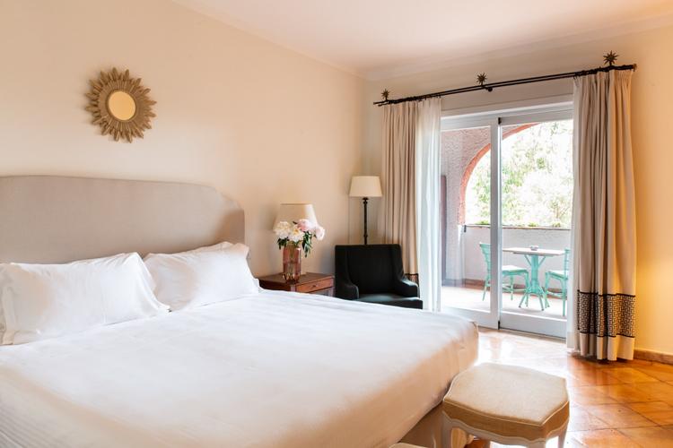 Crisp white linens and warm terracotta floors create an inviting sanctuary, while French doors frame glimpses of verdant gardens beyond the private balcony.