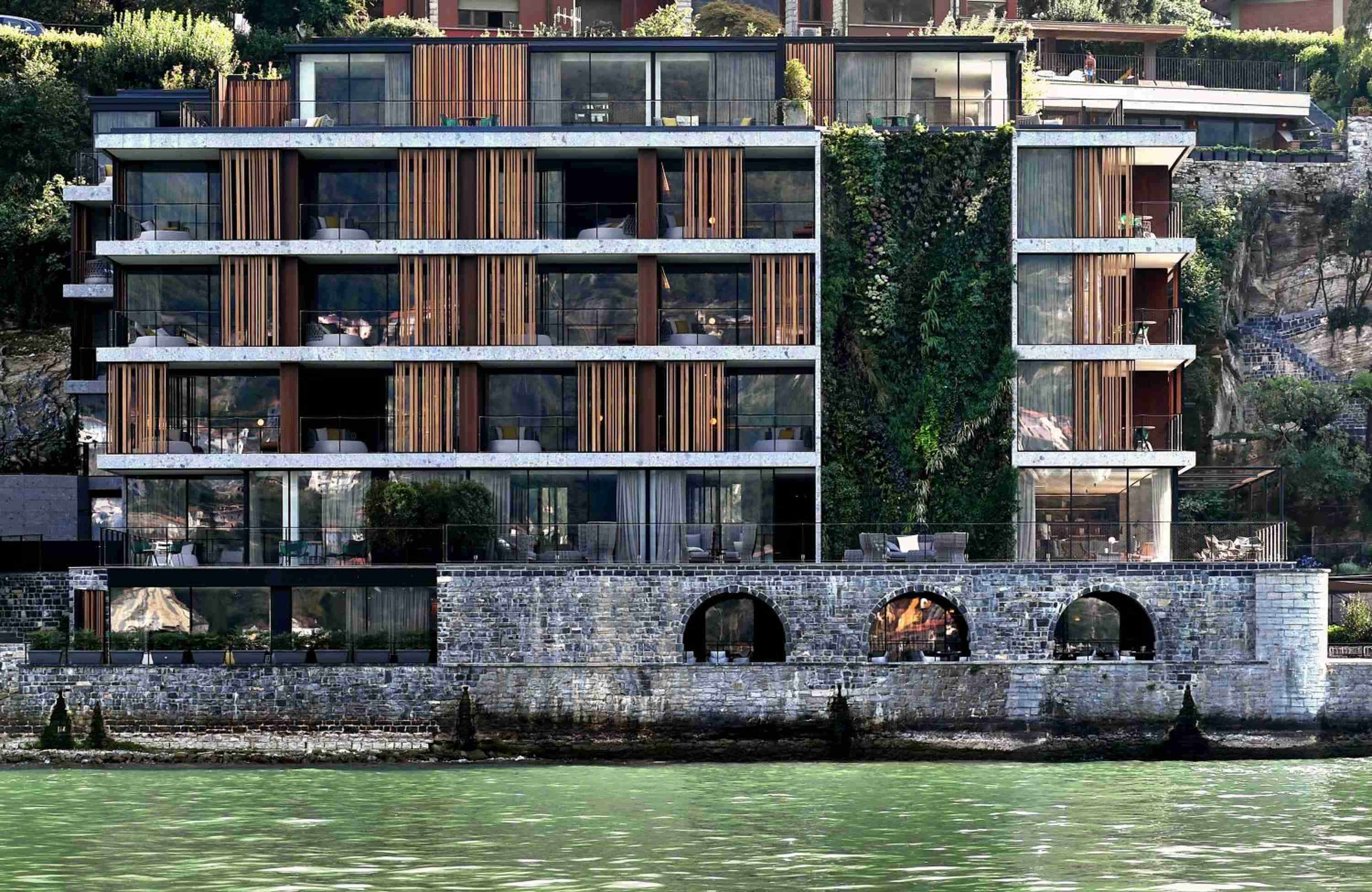 Modern glass and timber balconies cascade down the hillside above ancient stone arches, where Lake Como's emerald waters lap against centuries-old foundations.