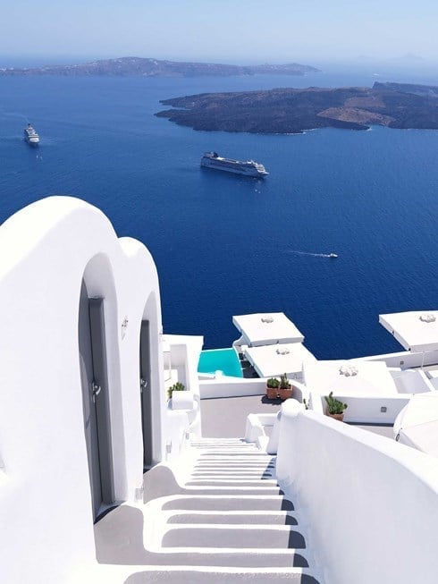 White-washed steps cascade down volcanic cliffs toward sapphire waters where cruise ships drift like toys beneath Santorini's endless azure sky.