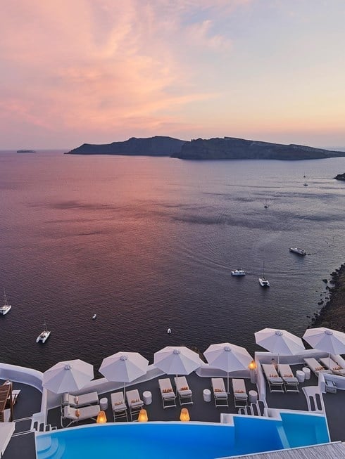 Infinity pool terrace melts into the Aegean's rose-gold embrace as sunset paints Santorini's caldera in ethereal pastels, white umbrellas standing sentinel against the twilight.