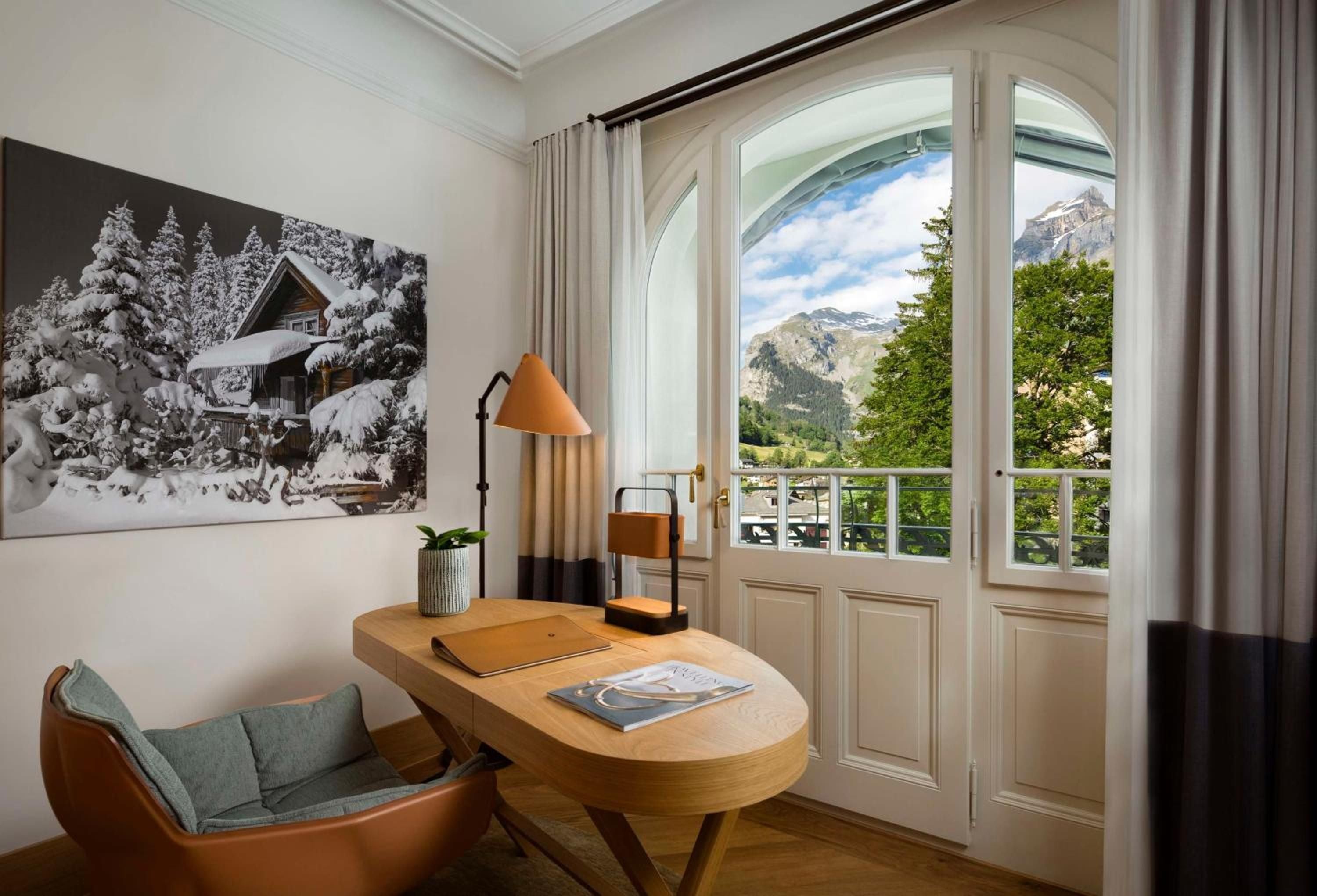 Warm wood furnishings and an orange task lamp create an inviting workspace, while arched windows frame breathtaking Alpine vistas beyond manicured gardens.