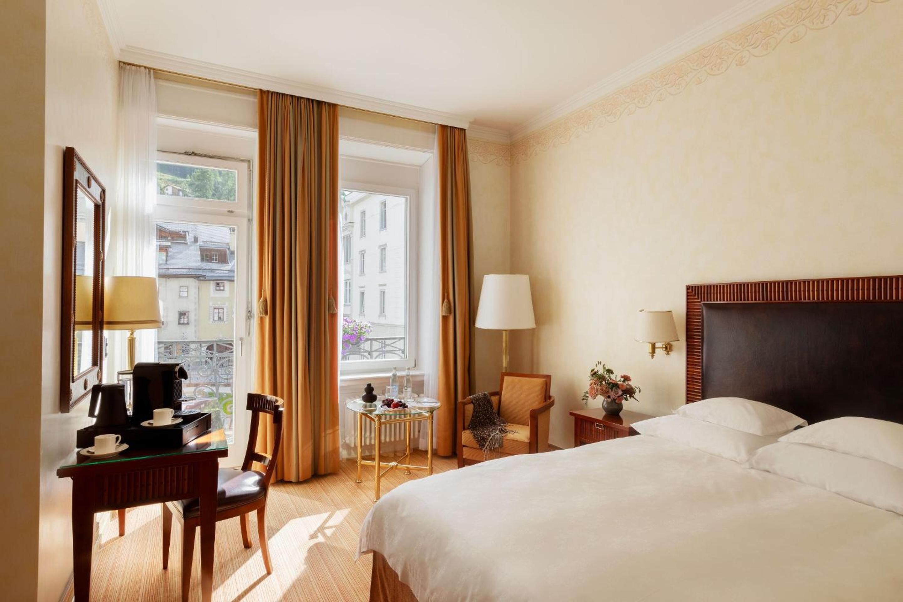 Sunlight streams through orange curtains into this elegant guest room, where polished wood furnishings and crisp linens frame alpine village views beyond French doors.