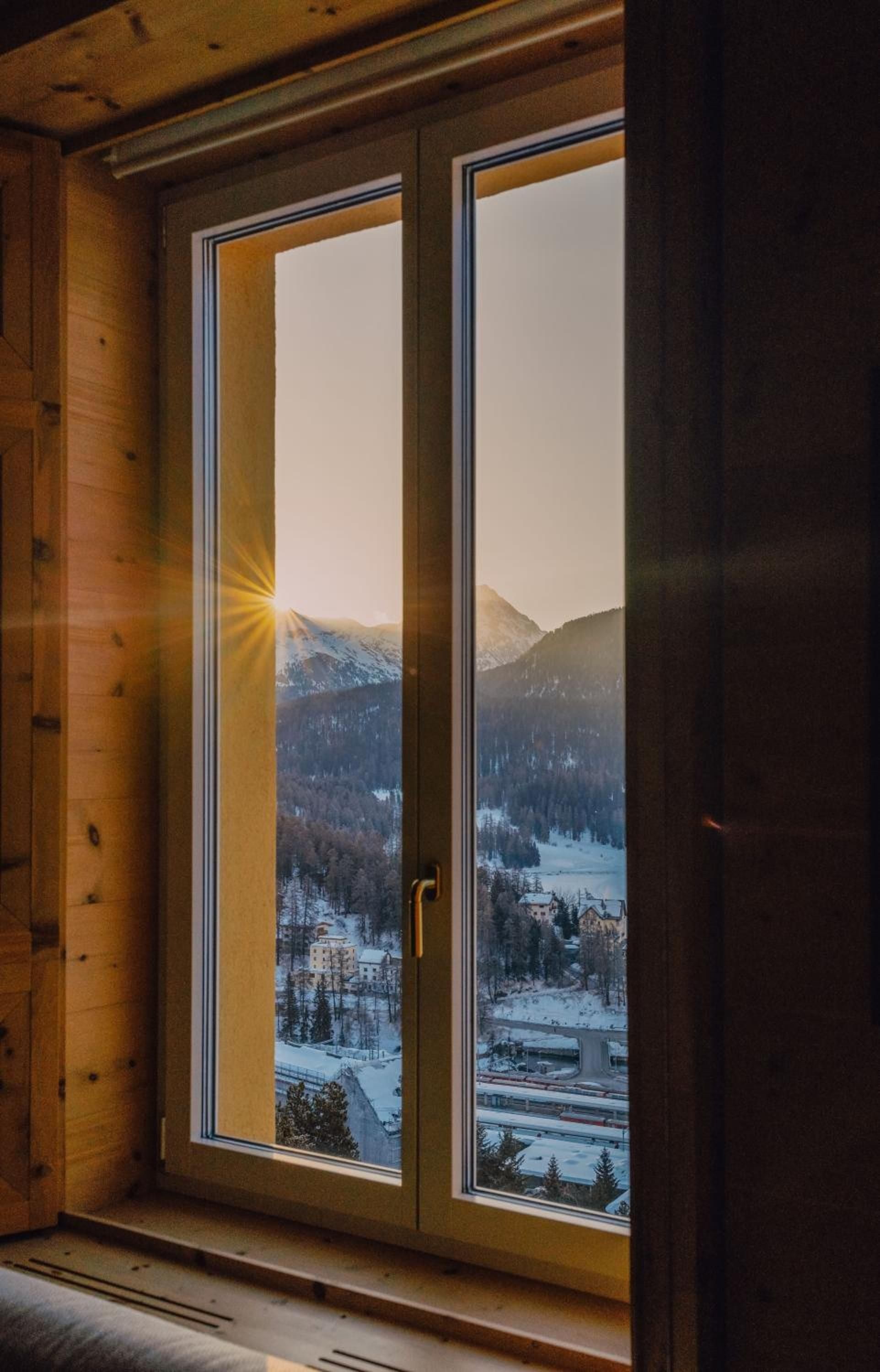 Sunburst pierces alpine peaks beyond hand-carved wooden window frames, where St. Moritz's snow-draped valley unfolds like a living postcard from your intimate mountain retreat.