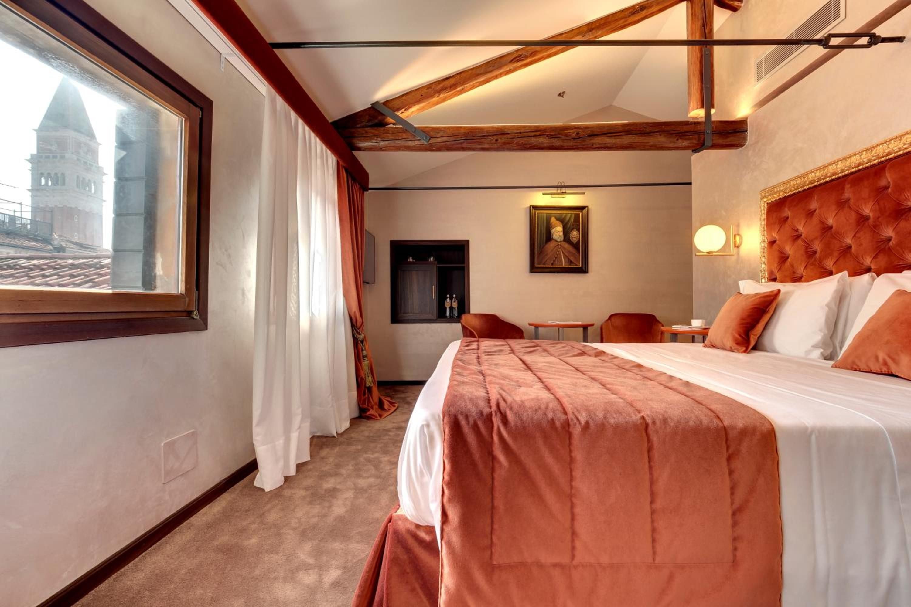 Exposed wooden beams frame views of Venice's bell towers from this elegant suite, where terracotta linens and period portraits create intimate luxury above the canals.