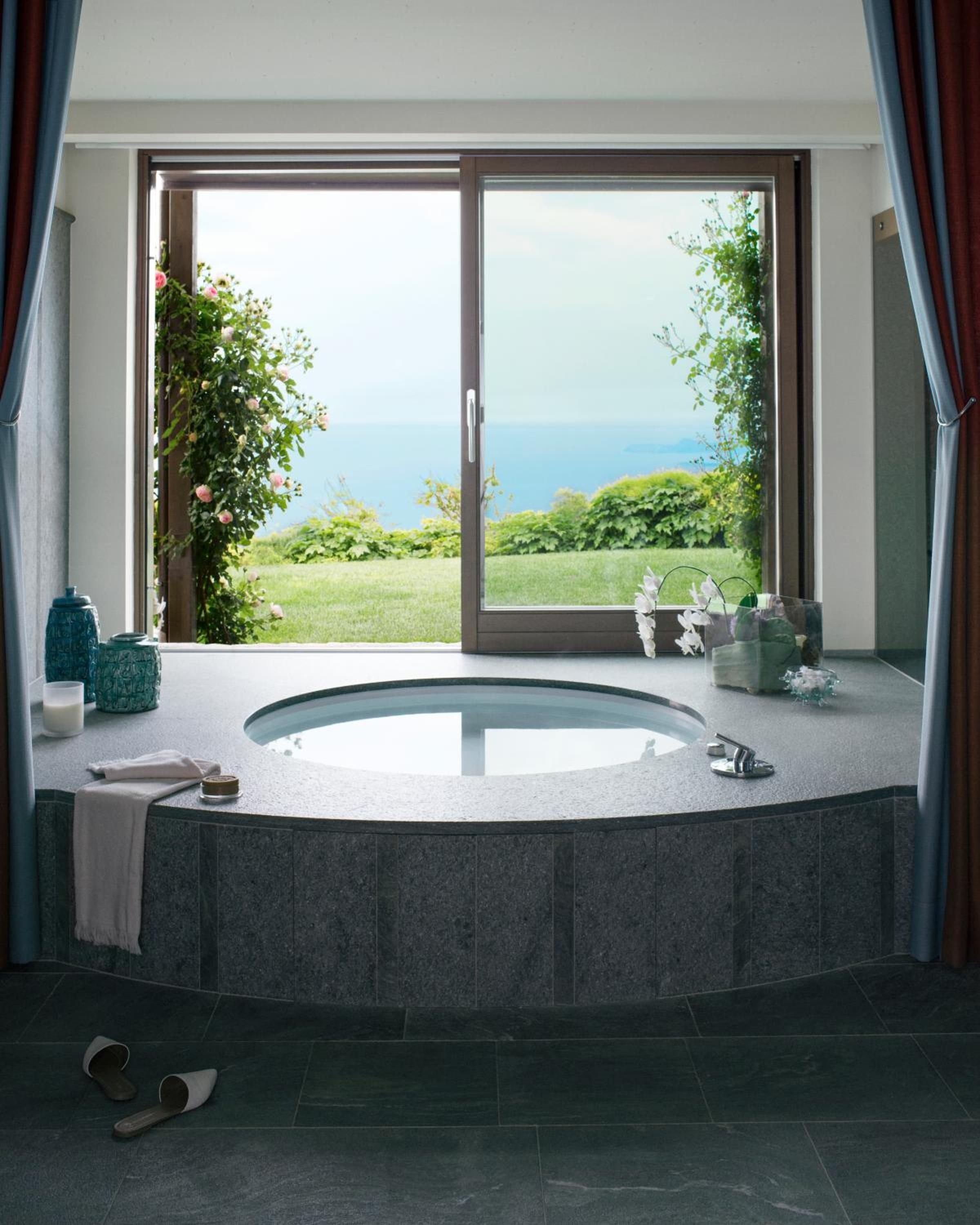 Stone-clad soaking tub frames Lake Garda's azure waters through floor-to-ceiling windows, where flowering vines and rolling lawns create an intimate spa sanctuary.