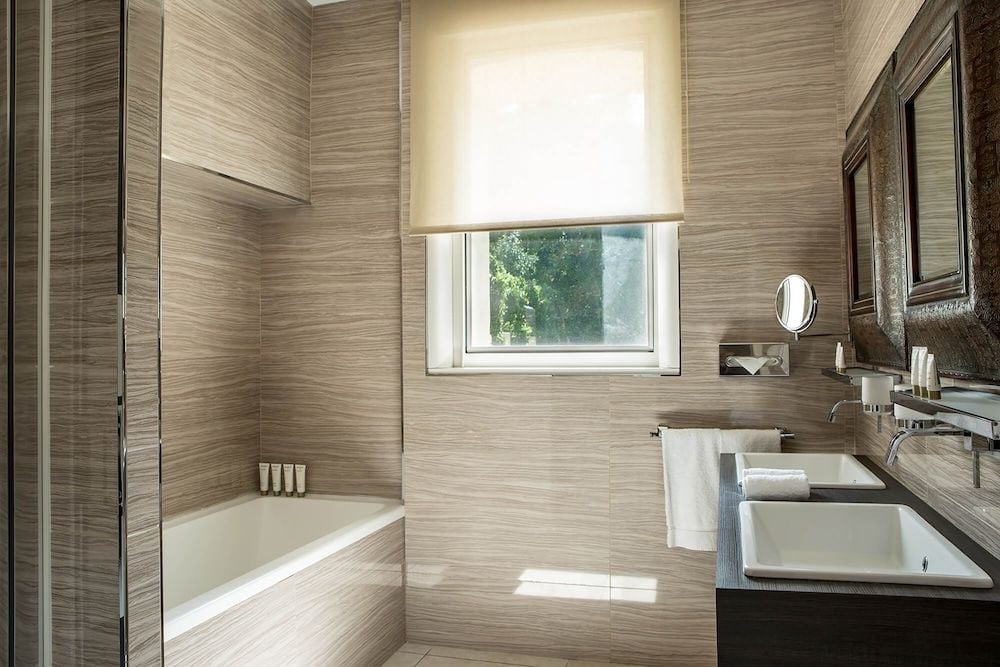 Natural light filters through bamboo shades onto warm wood-grain tiles, where dual sinks and a soaking tub create a spa-like sanctuary of understated luxury.