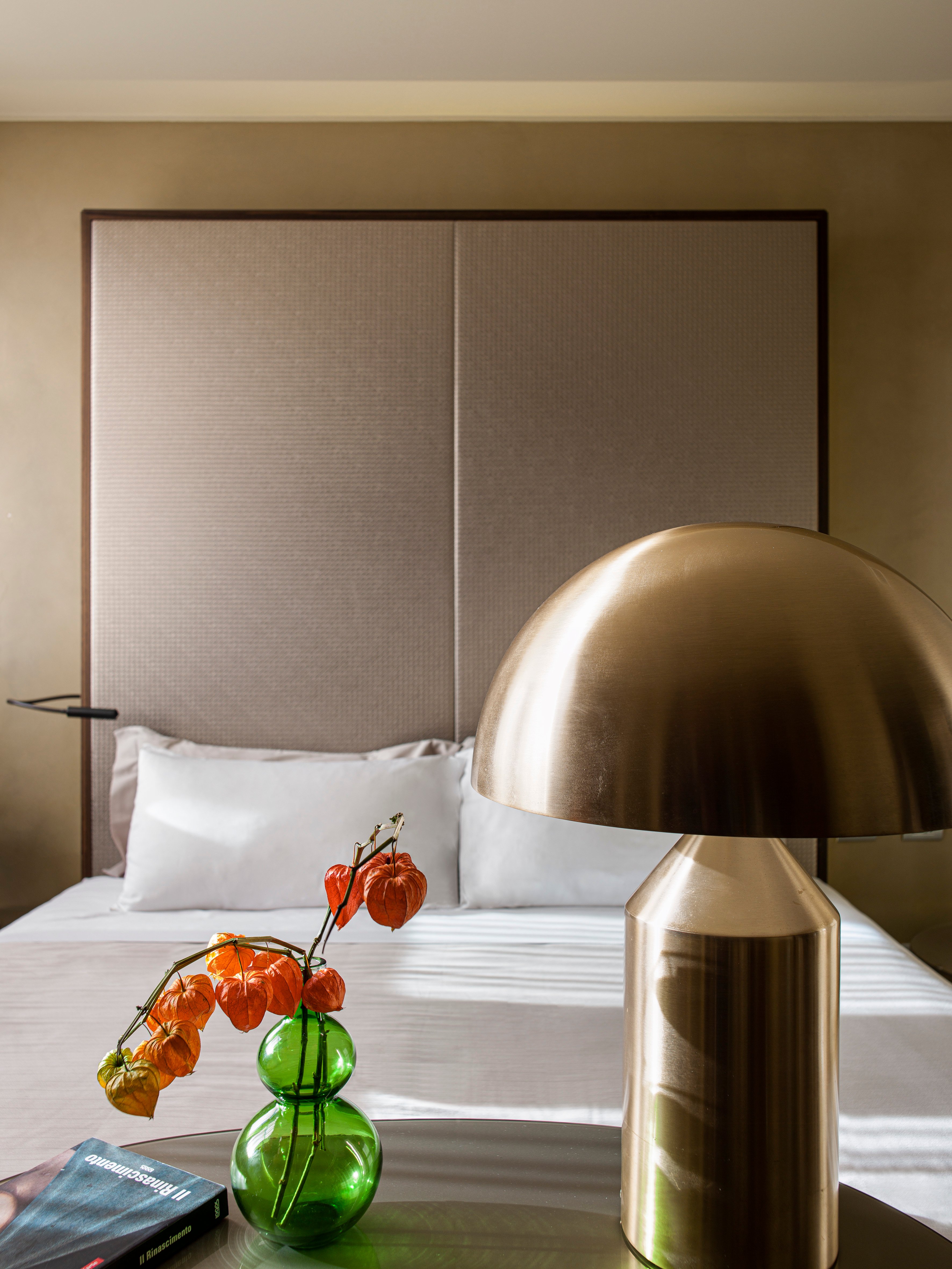 Warm brass lighting and autumn branches in emerald glass create an intimate vignette against the textured headboard, where contemporary Italian design meets seasonal elegance.