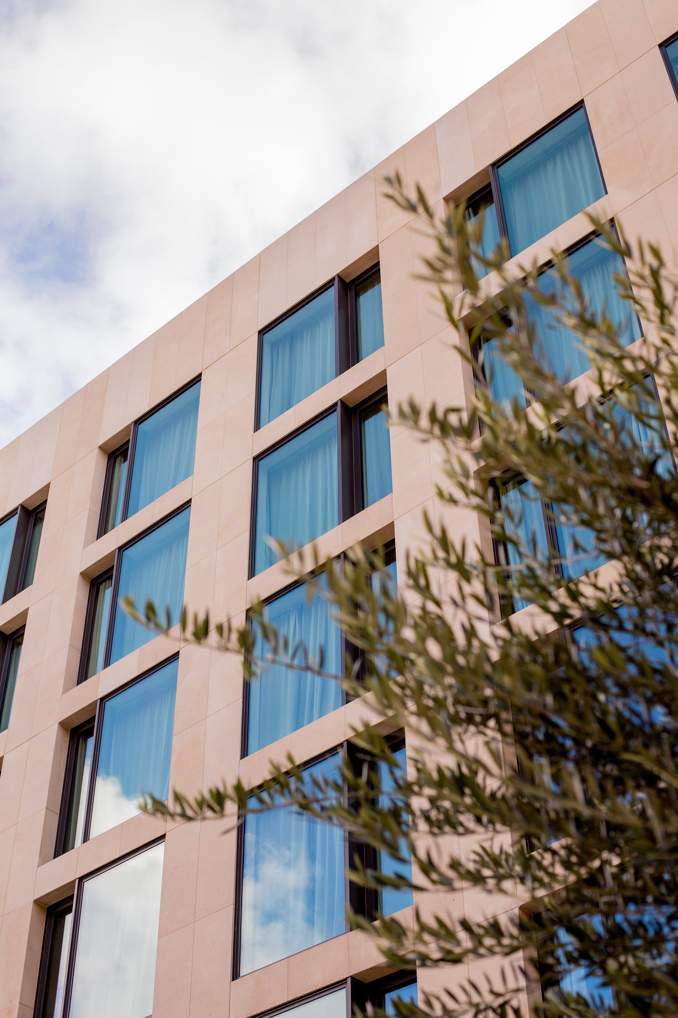 Sleek geometric windows punctuate ME Barcelona's coral-hued facade, where Mediterranean light dances across contemporary surfaces framed by native olive branches.