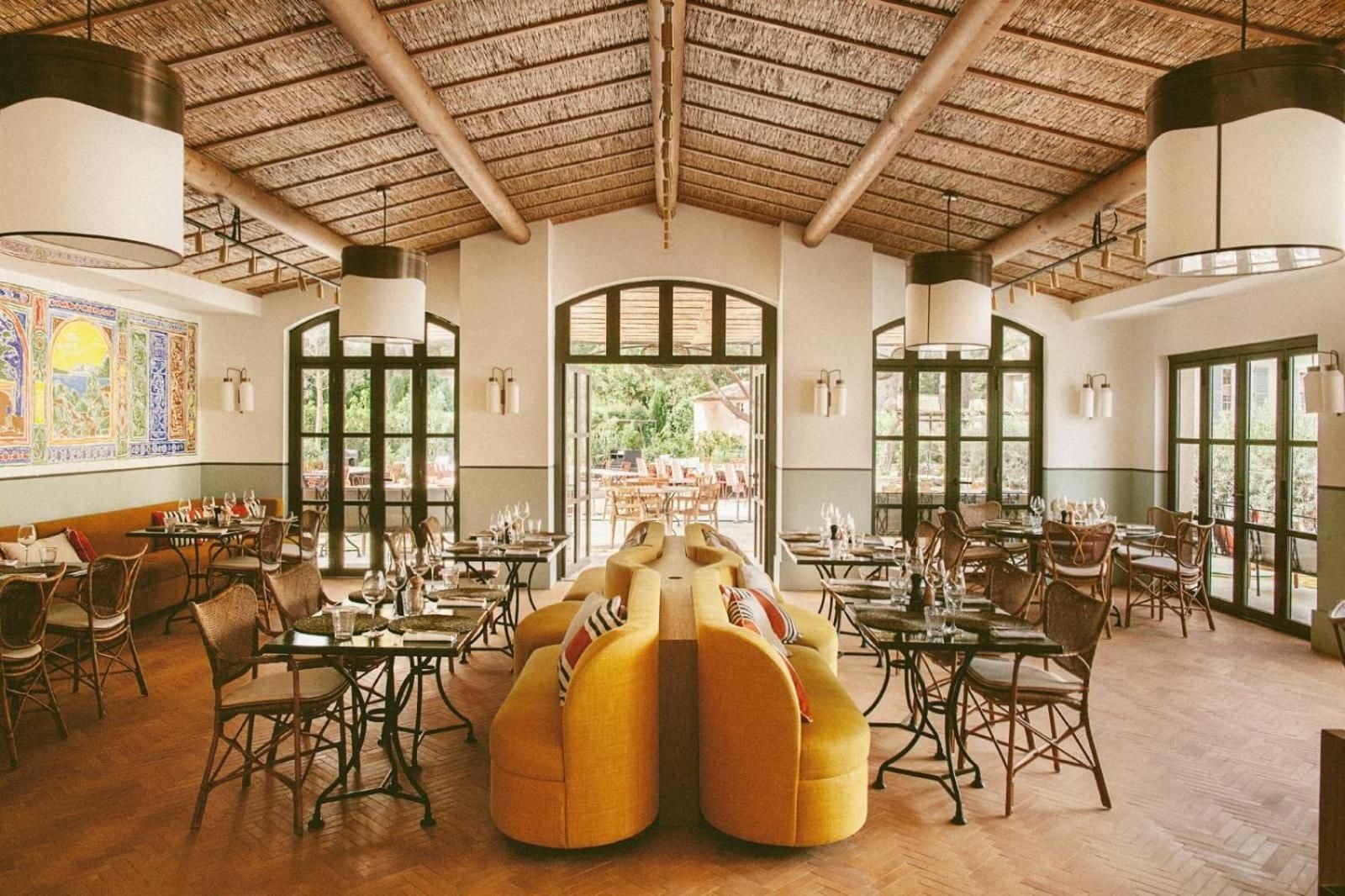 Woven reed ceilings and arched windows frame intimate dining tables, while golden banquettes anchor the sun-drenched Mediterranean restaurant that opens to verdant courtyards beyond.