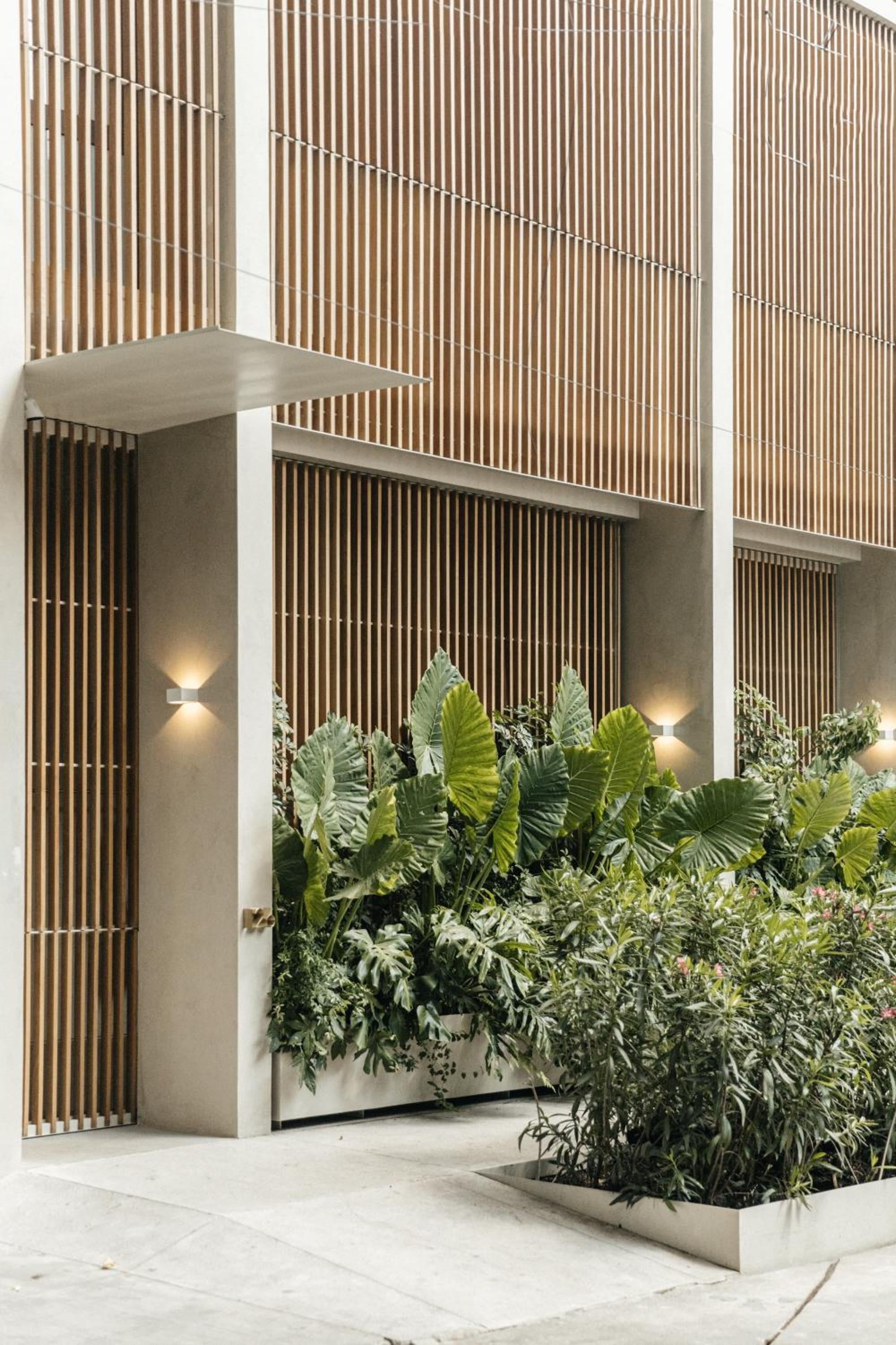 Vertical wooden slats create rhythmic shadows across concrete columns, while lush tropical plantings soften the contemporary entrance with warm, sculptural lighting.