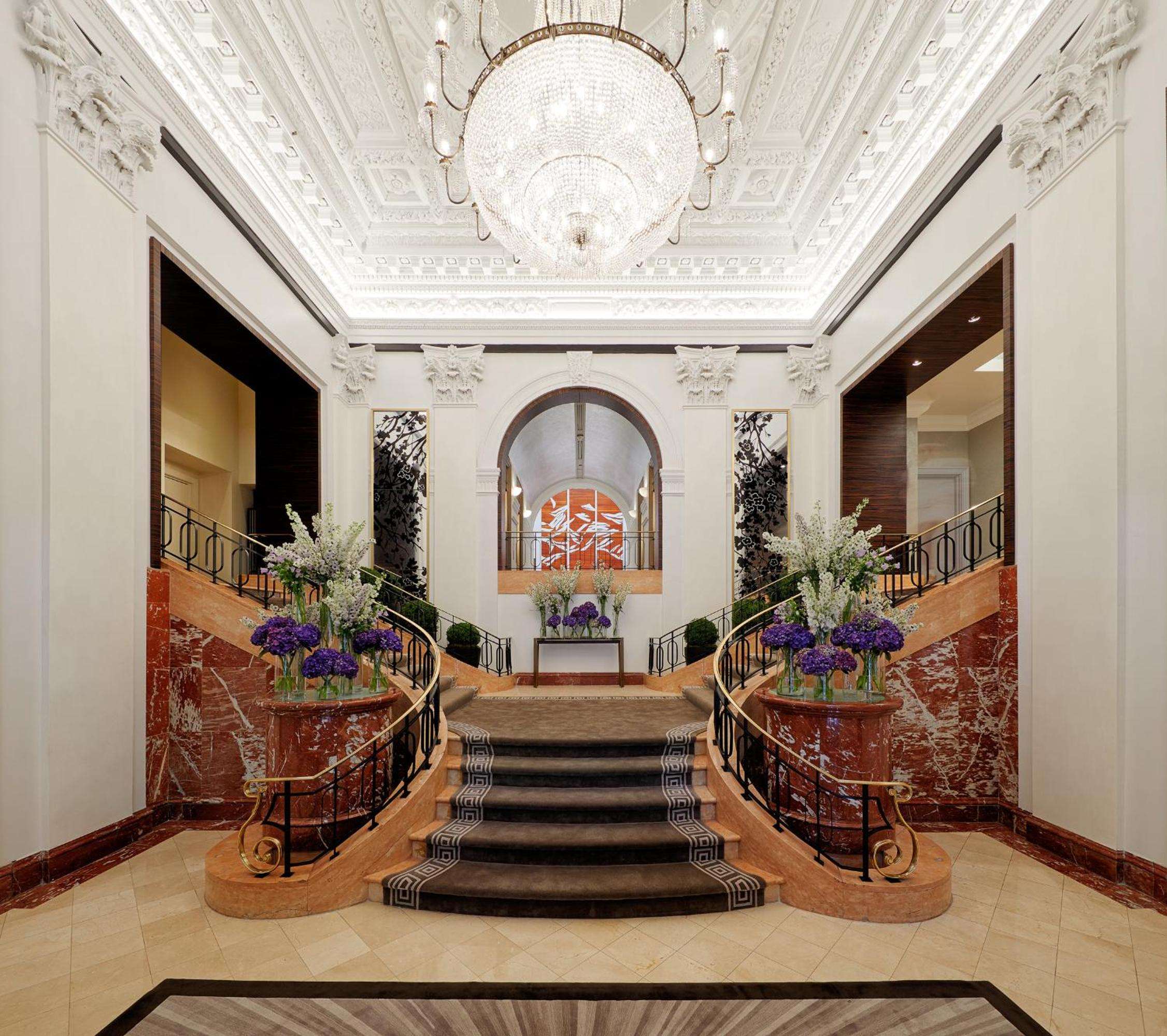 Ornate plasterwork crowns this grand staircase where crystal chandeliers cast warm light over marble balustrades and fresh hydrangeas, creating timeless Fifth Avenue elegance.