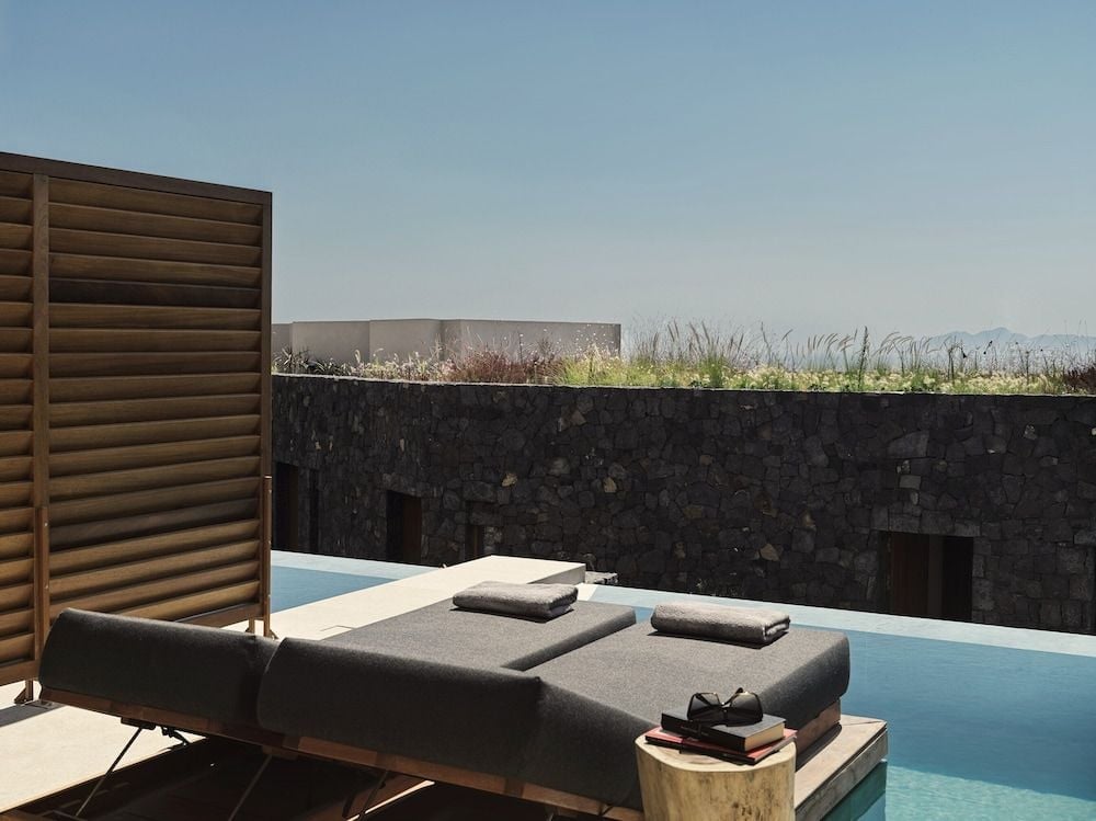 Dark stone walls contrast beautifully with azure waters, while wooden loungers invite afternoon languor beneath Santorini's endless sky and volcanic landscape.