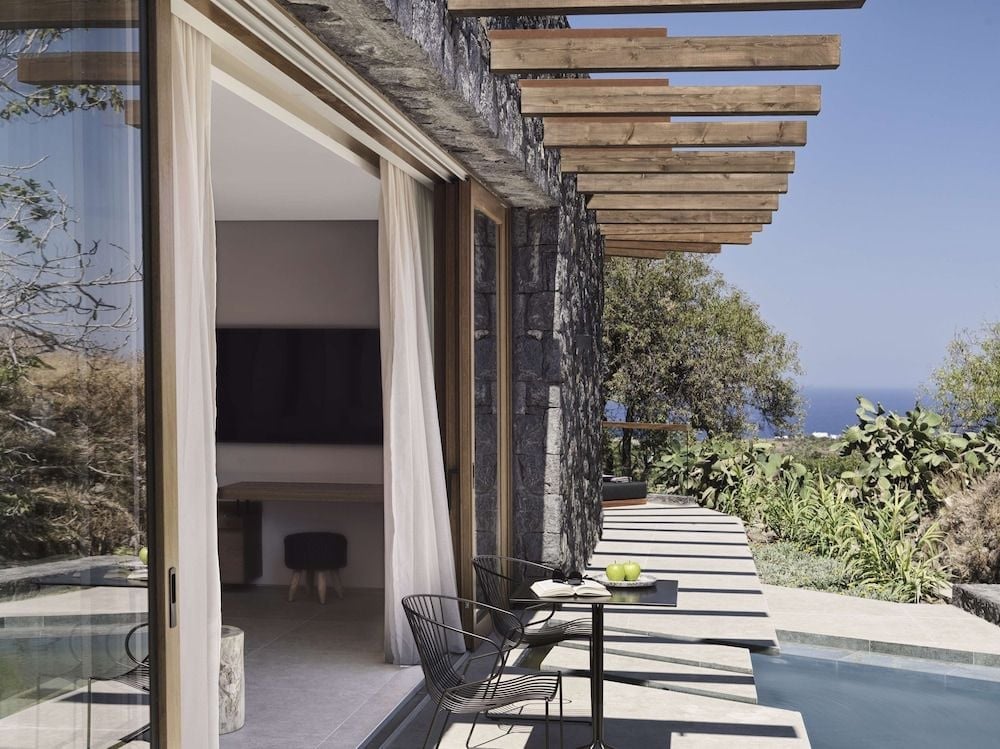 Stone walls and timber pergolas frame this poolside sanctuary, where flowing curtains catch Aegean breezes and volcanic rock meets modern minimalism in perfect harmony.