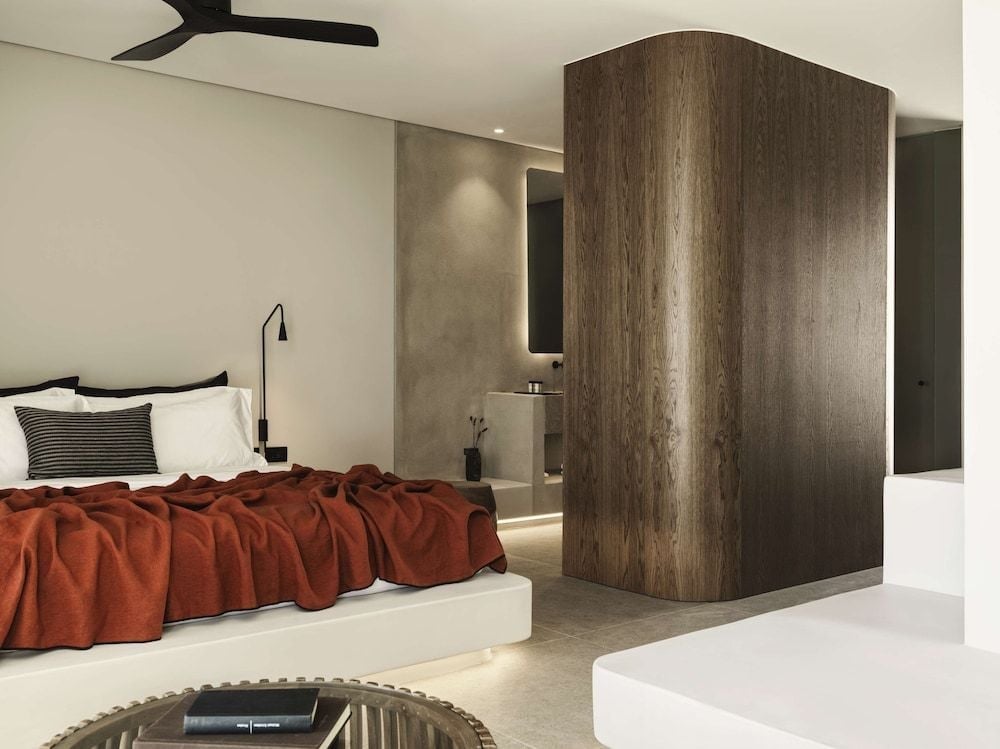 Rich terracotta linens contrast beautifully against minimalist white surfaces and warm wood paneling in this sophisticated suite, where understated luxury meets Cycladic simplicity.