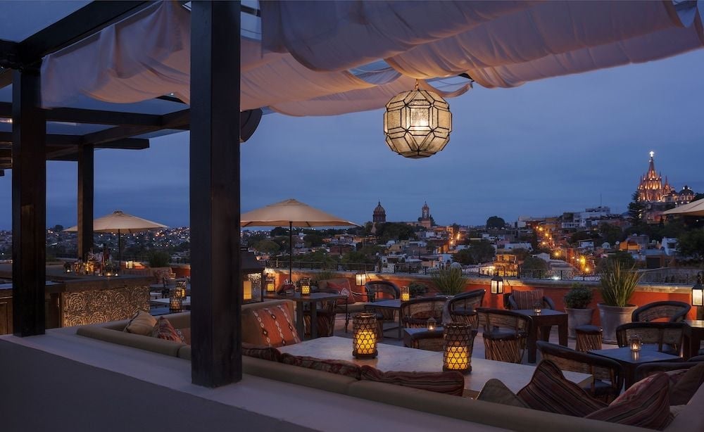 Golden lanterns cast warm pools of light across the rooftop terrace as San Miguel's iconic cathedral spires pierce the dusky sky beyond.