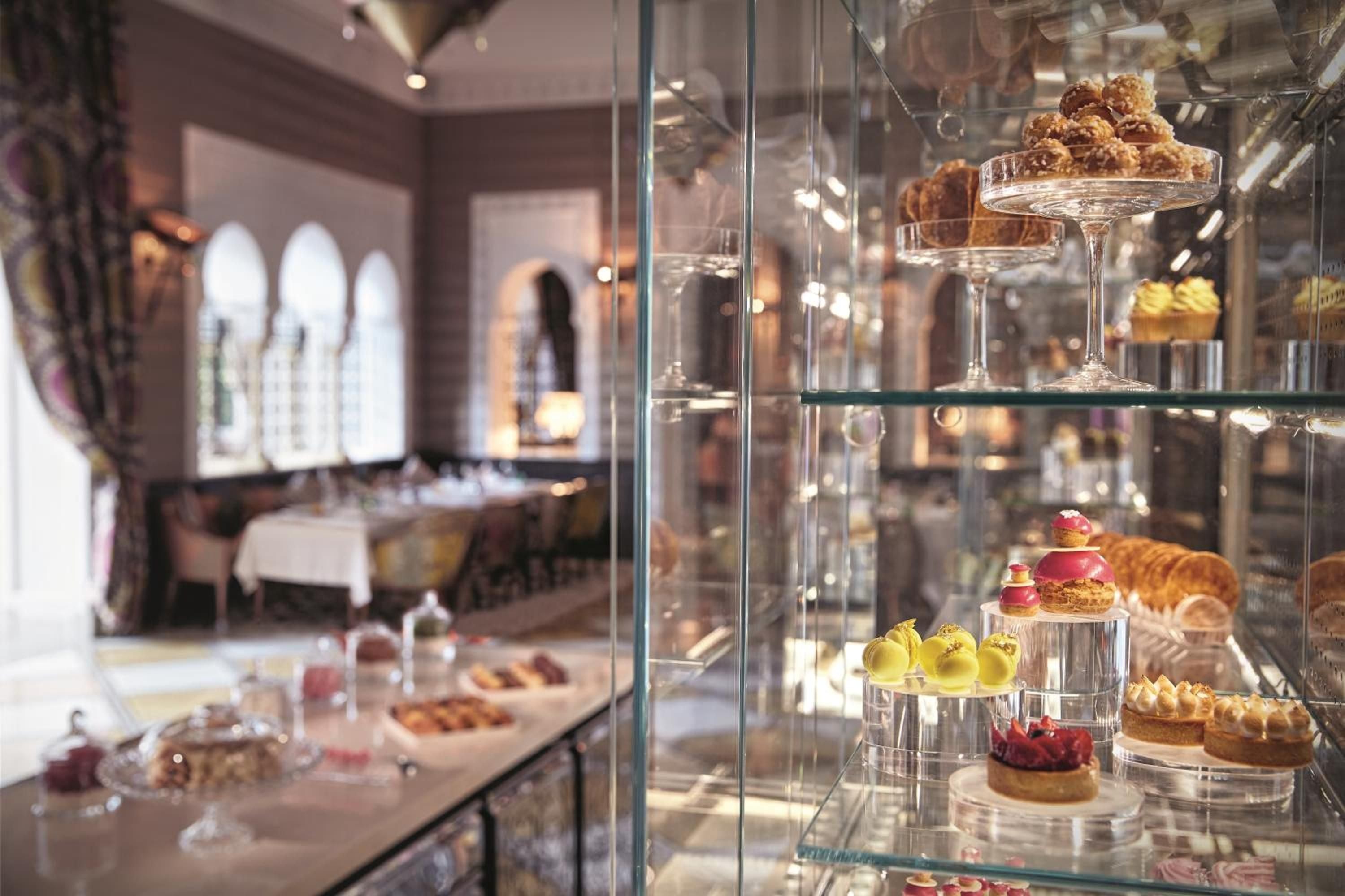 Exquisite pastries and colorful macarons gleam behind glass displays in the patisserie, where afternoon light catches crystal shelves and delicate French confections.