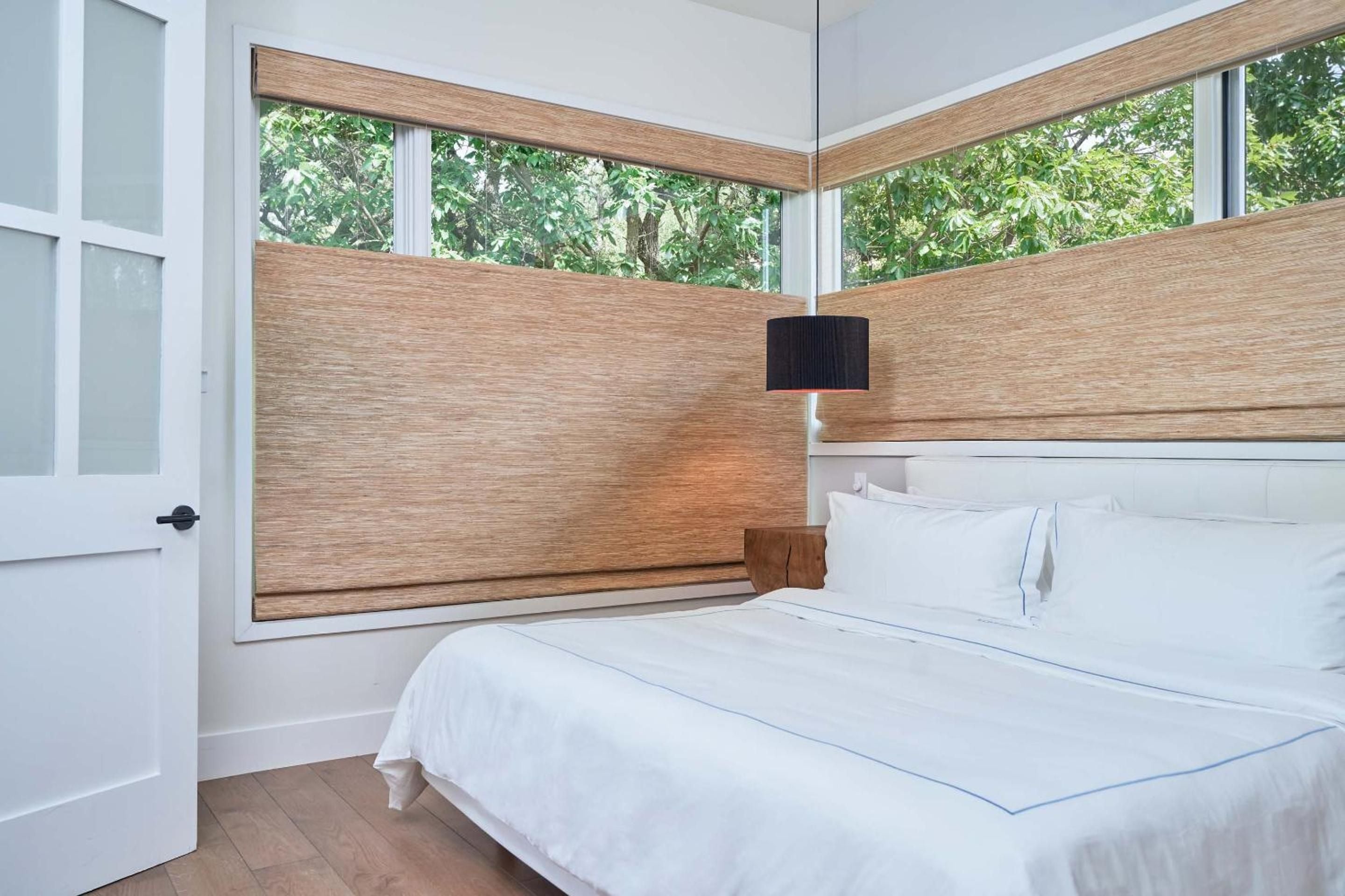 Corner windows frame verdant Austin canopy while natural wood privacy screens filter morning light across crisp white linens in this thoughtfully designed guest room.
