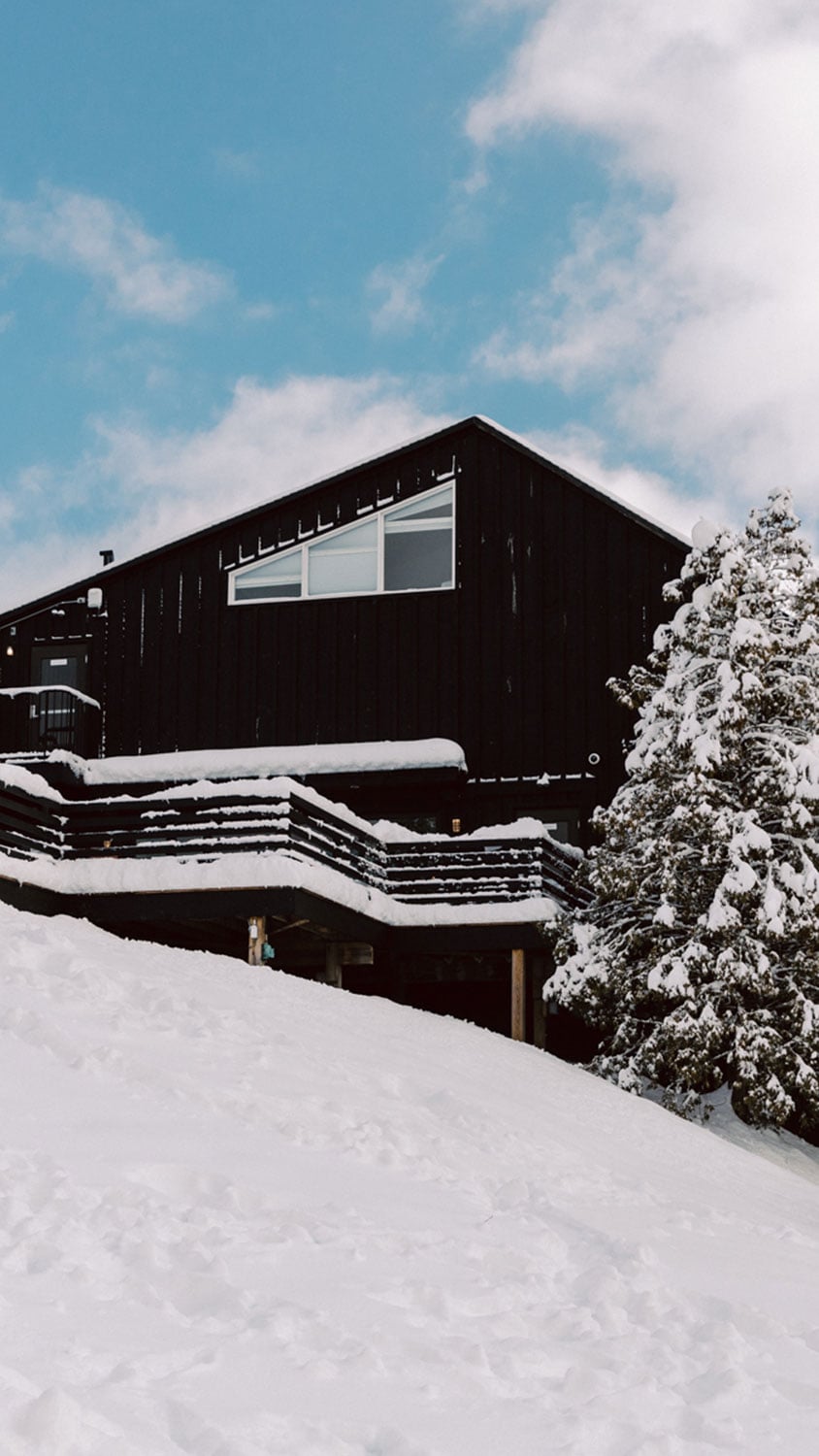 Snow blankets the lodge's dark timber facade and wraparound balconies, creating a striking contrast against powder-dusted evergreens and brilliant blue winter skies.